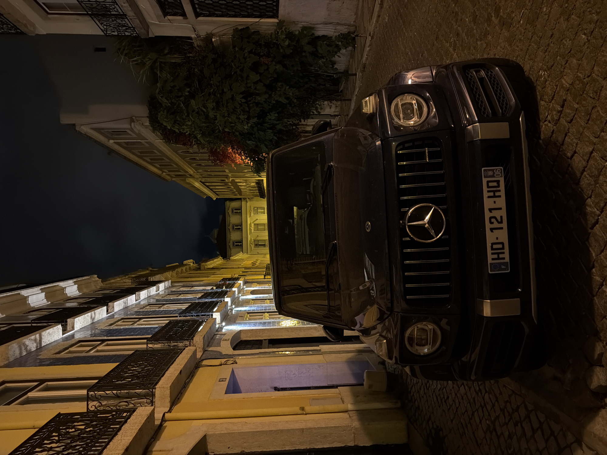Captured from an oblique perspective at night in Lisbon, Portugal, the scene features a black Mercedes-Benz G-Class SUV dominating the right side of the frame. The vehicle's front grill and license plate, "HD-121-HQ," are visible. The street, paved with cobblestones, is narrow, flanked by aged buildings with wrought-iron balconies. These structures are illuminated by warm, artificial light, emphasizing their architectural details. Vegetation, including what appears to be a vine, grows along one of the building's facades. The sky above is dark, hinting at the late hour. No people are visible in the frame.