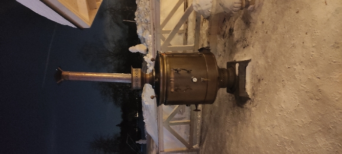 An outdoor, brass-colored samovar sits on a snowy surface. The samovar features a cylindrical body with a chimney rising from the top. A temperature gauge is visible on the body. The background includes a dark sky, trees, a white fence, and a building corner. The samovar appears used, with visible wear and possible soot stains.