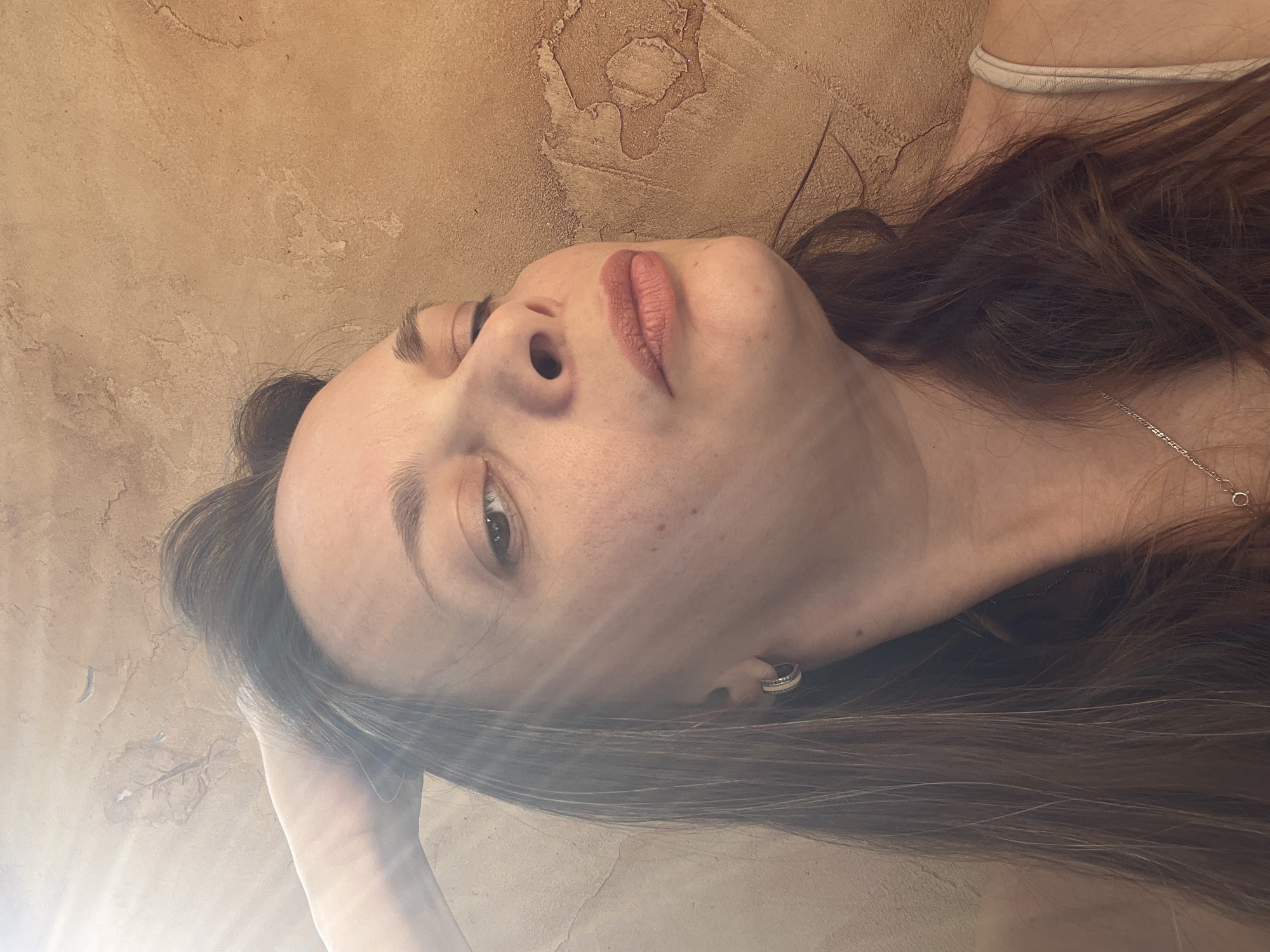 A close-up, low-angle, waist-up shot shows a woman with medium-length brown hair and light skin. She is looking upwards with her left hand raised to her head. She is wearing a light-colored tank top and a silver necklace. She has a hoop earring in her visible left ear. The background is a textured, tan wall. Sunlight beams in from the upper left. The image was taken in Tolyatti, Russia.