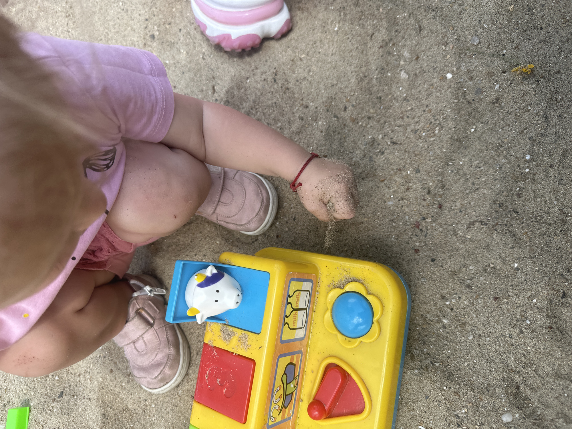 A child in a pink outfit and pink shoes is playing with a yellow toy in a sandpit. The toy has buttons and images of farm animals and milk bottles. The child's hands and legs are covered in sand. The scene appears to be outdoors, likely in a park or playground, given the presence of a sandpit. The context of Donchovci, Bulgaria, suggests this is a local scene.