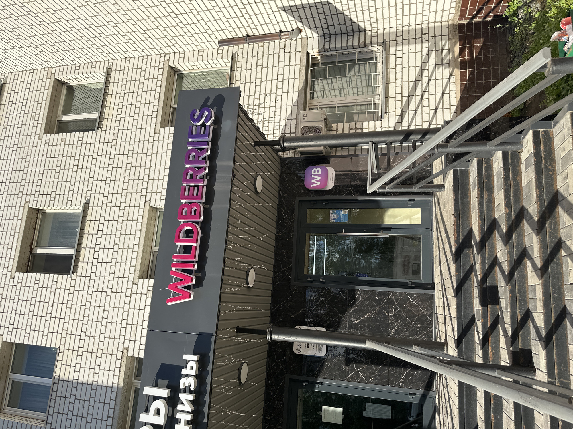 The image shows the entrance to a Wildberries store in the city of Rubtsovsk, Russia. The store is located on the ground floor of a white brick building. The entrance features a dark marble facade with a prominent "WILDERRIES" sign in pink and purple lettering. To the left of the entrance, a portion of another sign is visible, with white letters on a dark background. A set of concrete stairs with metal railings leads up to the entrance. A small white sign with a purple "WB" logo is mounted on the facade to the left of the door. An air conditioning unit is visible on the wall above the entrance. The scene is illuminated by sunlight, casting strong shadows on the stairs and the building's facade. There are no people visible in the image.