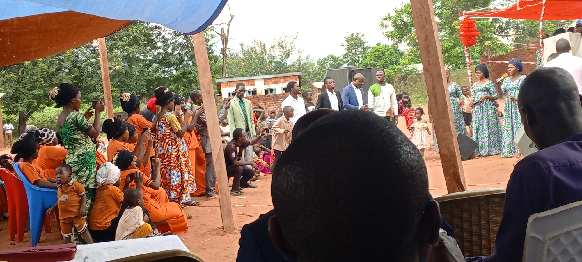A medium shot captures an outdoor gathering in Ngalula, Democratic Republic of the Congo. A crowd of people is present, many dressed in vibrant orange and green patterns. Some individuals are holding and using mobile phones to record the event. Several children are also in attendance. A group of men in suits stands near the center, while women in matching green and blue patterned dresses stand in front of a microphone, possibly performing or speaking. Overhead, a blue and brown tarp provides partial shade. A simple brick building can be seen in the background, along with trees. The scene appears to be a cultural or community event.