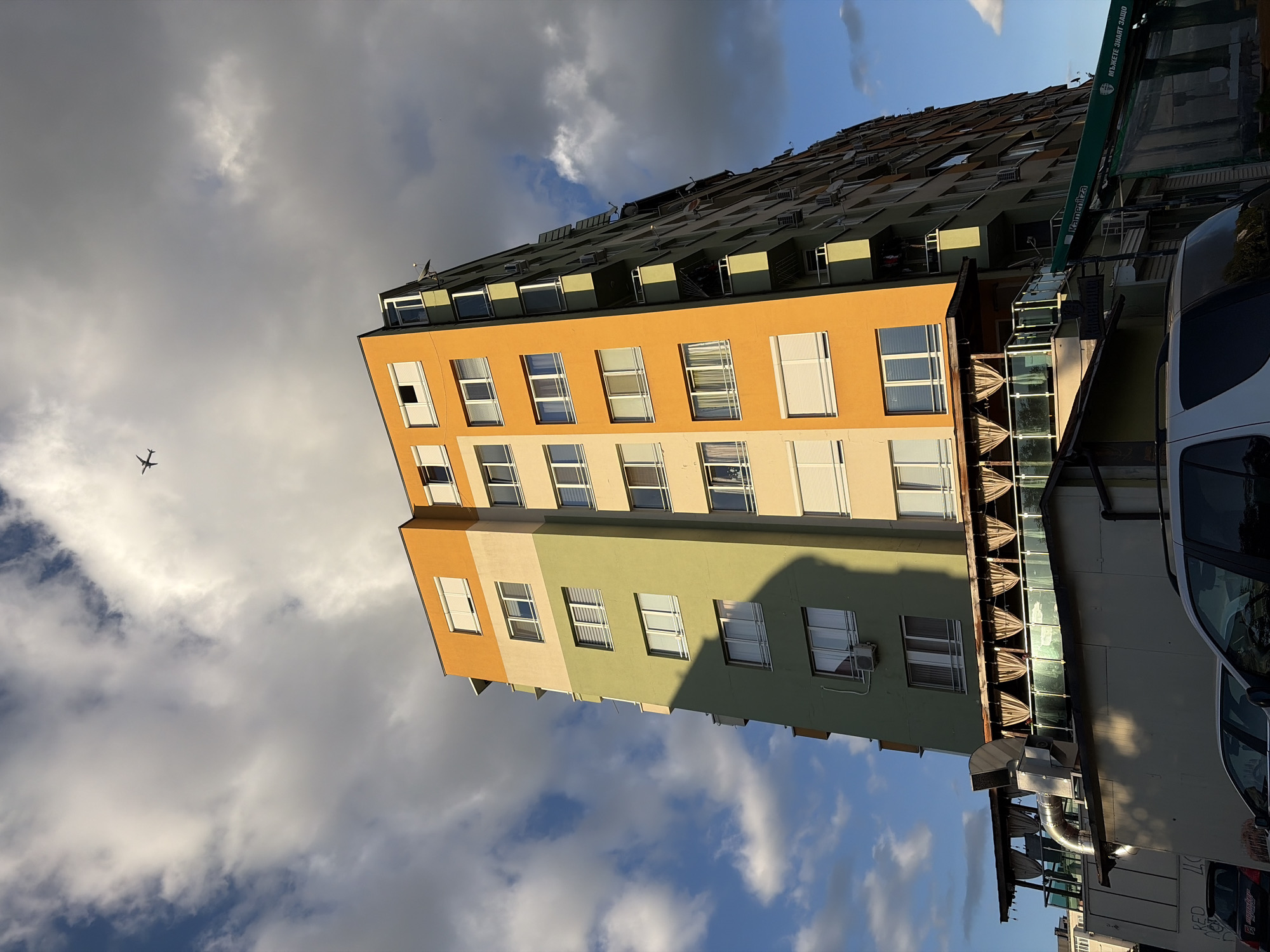 A low-angle, full shot captures a colorful apartment building under a dynamic sky with clouds and a distant airplane. The building has sections painted orange, light beige, and light green, stacked vertically. Numerous windows, some with white blinds, dot the facade. A small airplane is visible high in the sky, silhouetted against the clouds. Below the apartment building, a covered outdoor seating area with tables and chairs is visible, protected by curtains. Several cars are parked in front of the building. The lighting suggests it is late afternoon or early evening, with strong shadows cast by the building onto itself and the foreground.