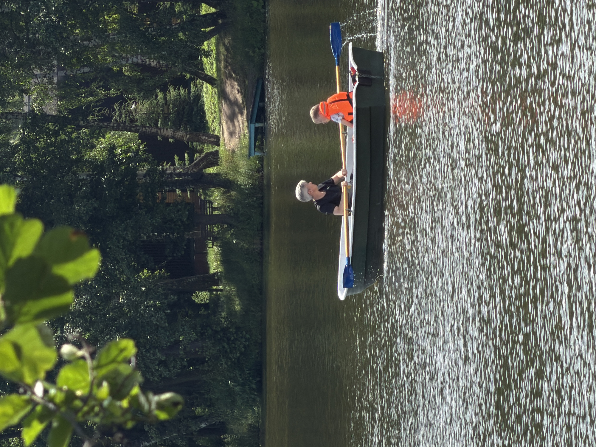 In Malakhovka, Russia, on a body of water, a woman with short, gray hair, wearing a black t-shirt, sits in a small, dark green rowboat. Facing her, a young child, likely a boy, is seated and wears an orange life vest. The woman holds a blue and yellow oar, appearing to row the boat, as indicated by the splashing water behind it. The surrounding environment is lush with trees and vegetation, suggesting a natural setting within the city. The water's surface is rippled, reflecting the sunlight. The scene captures a moment of leisure and outdoor activity.