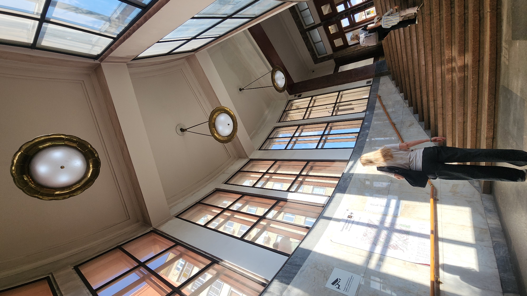 A wide-angle, low-angle shot captures the interior of a grand staircase in Moscow, Russia, bathed in sunlight streaming through large, multi-paned windows. A woman with blonde hair, wearing a light-colored t-shirt and dark trousers, stands in the foreground, facing away from the camera and towards the stairs. She carries a black shoulder bag. Behind her, a set of wide, red-brown stone stairs ascends diagonally to the right. Further up the stairs, two other individuals are visible: a person wearing a backpack and dark clothing, and a child in a striped shirt and light-colored trousers walking upwards. The walls are adorned with patterned marble on the lower section and a large informational map on the left. The ceiling features ornate recessed panels and two large, circular brass pendant lights with white diffusers, identical to one prominently displayed in the upper left corner of the frame. The windows reflect the facades of buildings.