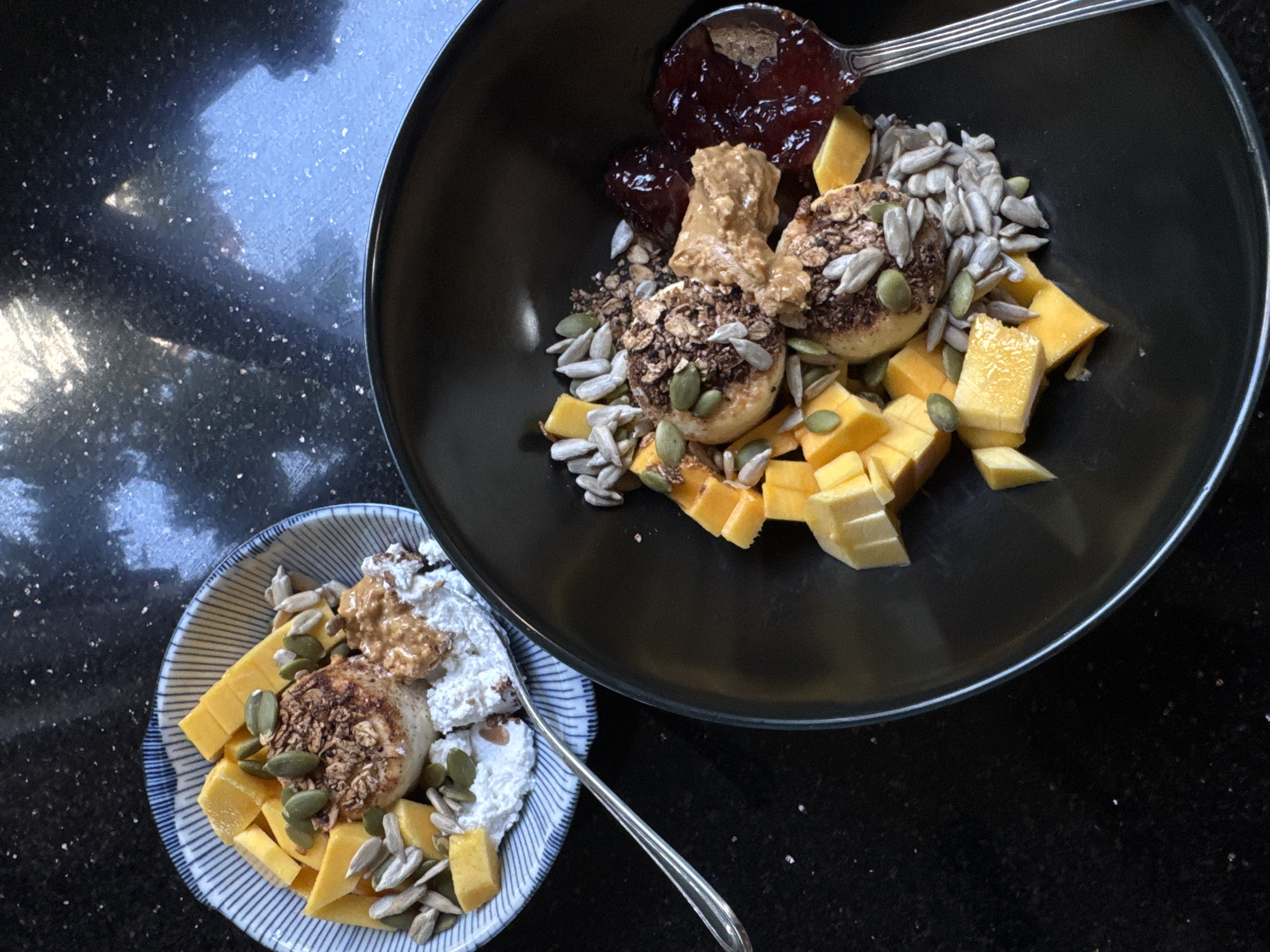 Two bowls of breakfast are presented on a speckled black countertop. The bowl on the left is white with blue stripes and contains cottage cheese, cubed mango, granola, pumpkin seeds, sunflower seeds, and a dollop of peanut butter. A fork rests to the left of this bowl. The bowl on the right is black and filled with cottage cheese, cubed mango, granola, pumpkin seeds, sunflower seeds, peanut butter, and a spoonful of dark red jam. A spoon is positioned to the right of this bowl. The scene is captured from a top-down perspective, with light reflecting off the countertop, creating a glossy effect. No individuals or specific landmarks of Goviyapana, Sri Lanka, are visible in the image.