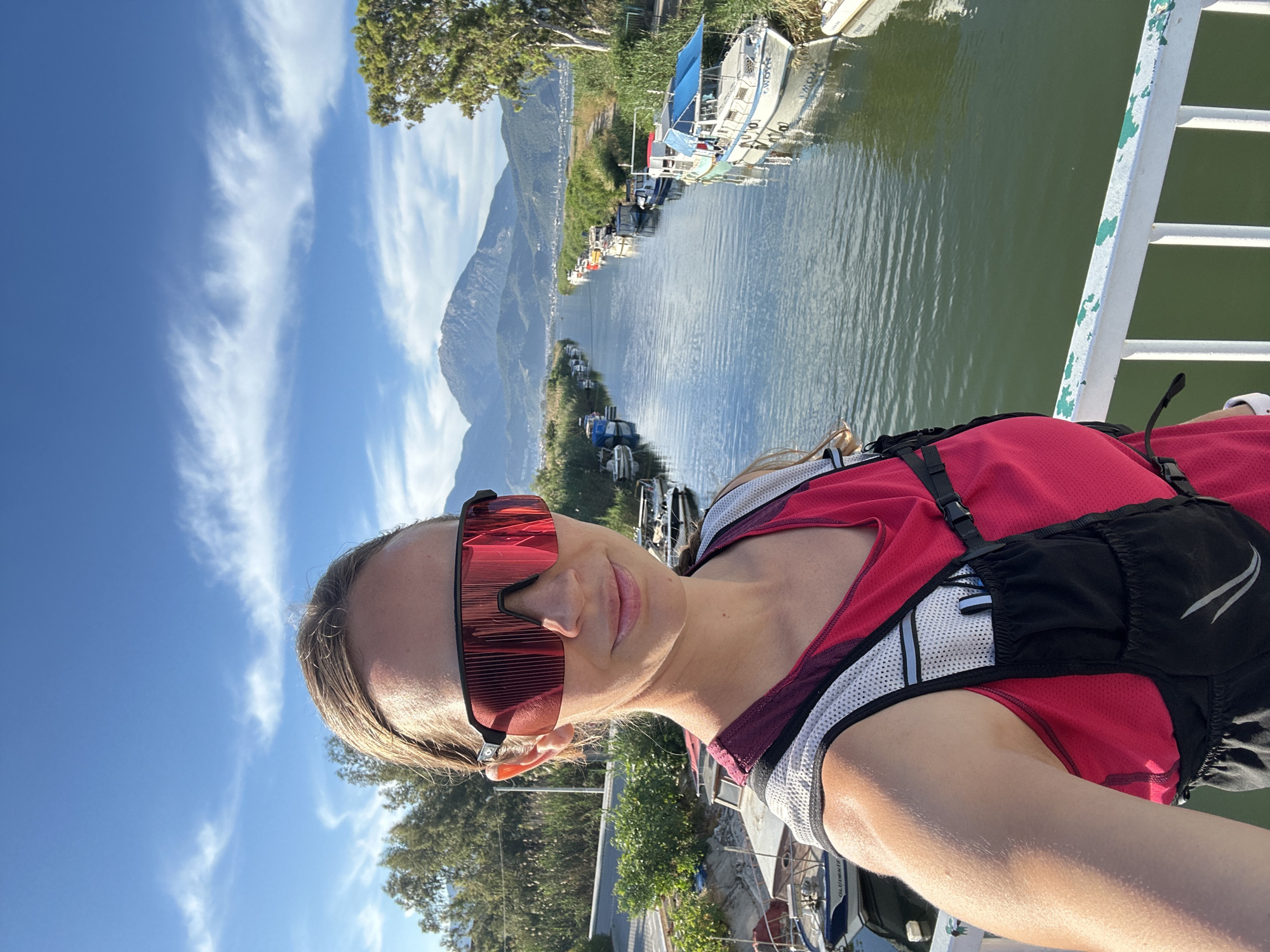 A woman stands on a bridge overlooking a river or canal in Fethiye, Turkey. She is wearing a red and white top, a black hydration pack, and red sunglasses. The water reflects the sky and the nearby mountains. Boats are docked along the river's edge. The sky is blue with scattered clouds. The bridge appears to be made of metal and painted white. Vegetation lines the riverbanks.
