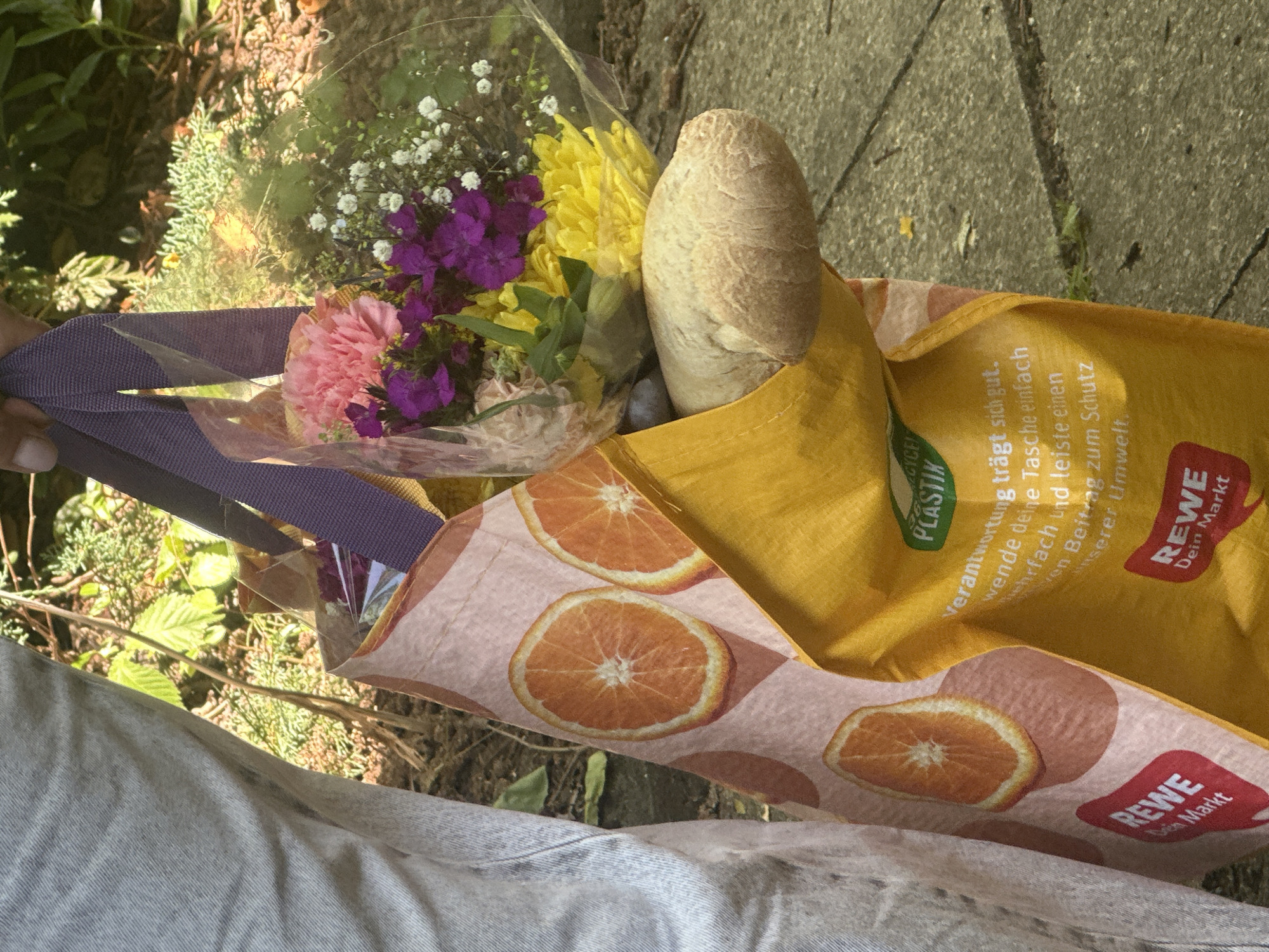 A person's hand holds a reusable shopping bag filled with a bouquet of flowers and a baguette. The bag is yellow with an orange slice pattern and features text in German, including "REWE Dein Markt" and "Verantwortung trägt sich gut. ... wende deine Tasche einfach mehrfach und leiste einen Sven Beitrag zum Schutz unserer Umwelt." The bouquet includes pink carnations, purple flowers, and small white flowers, wrapped in clear plastic. The scene appears to be outdoors, with a concrete path visible in the background and foliage and soil to the left, suggesting a daytime setting. The image is taken from a slightly elevated angle, looking down into the shopping bag. There are no discernible emotional cues or specific time of day indicators beyond the natural lighting.