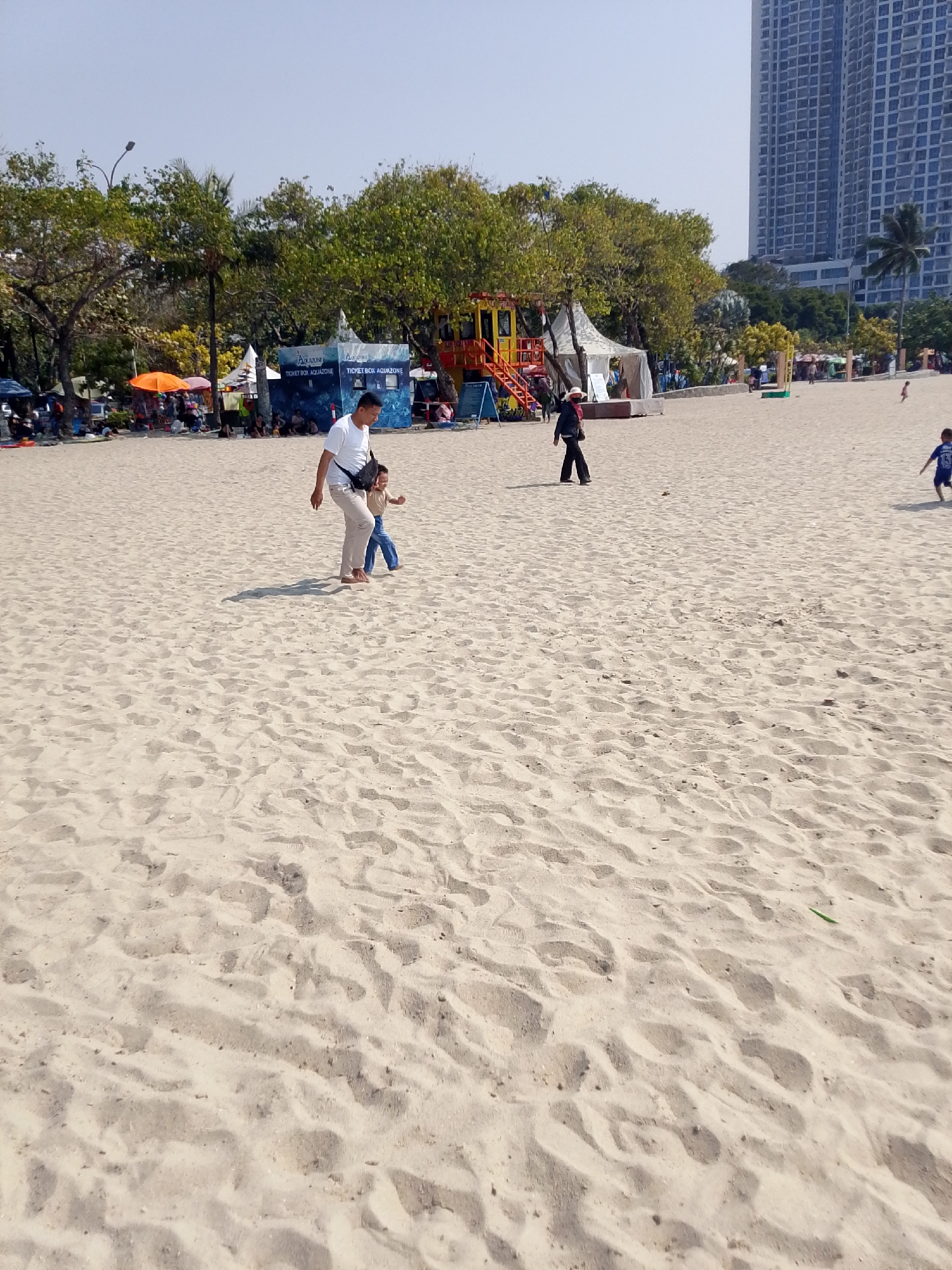 A daytime, wide-angle, eye-level shot captures a sandy beach with a backdrop of trees, tents, a playground structure, and a tall building. In the foreground, a man in a white t-shirt and tan pants walks with a young child dressed in a blue shirt and pants. The man has a black bag slung across his chest. Behind them, a woman in black pants and a black top walks with her head down. Further back, on the left, a line of tents and stalls is visible, with people seated or standing around them. An orange umbrella is prominent among the stalls. To the right and in the background, a large, modern, multi-story building dominates the skyline, with palm trees and other foliage scattered around the beach area. The sand is textured with numerous footprints and slight undulations.