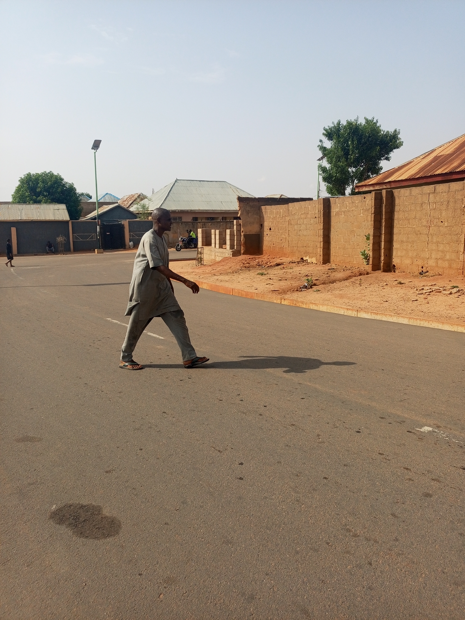An individual dressed in gray clothing is walking across a paved road. To the right, there is a tan brick wall and patches of red dirt. In the background are buildings with corrugated metal roofs and trees. A street light is also visible. Other individuals and a motorbike can be seen in the distance on the left. A stain is present on the road in the foreground. It is daytime. The city is Tempure, Nigeria.