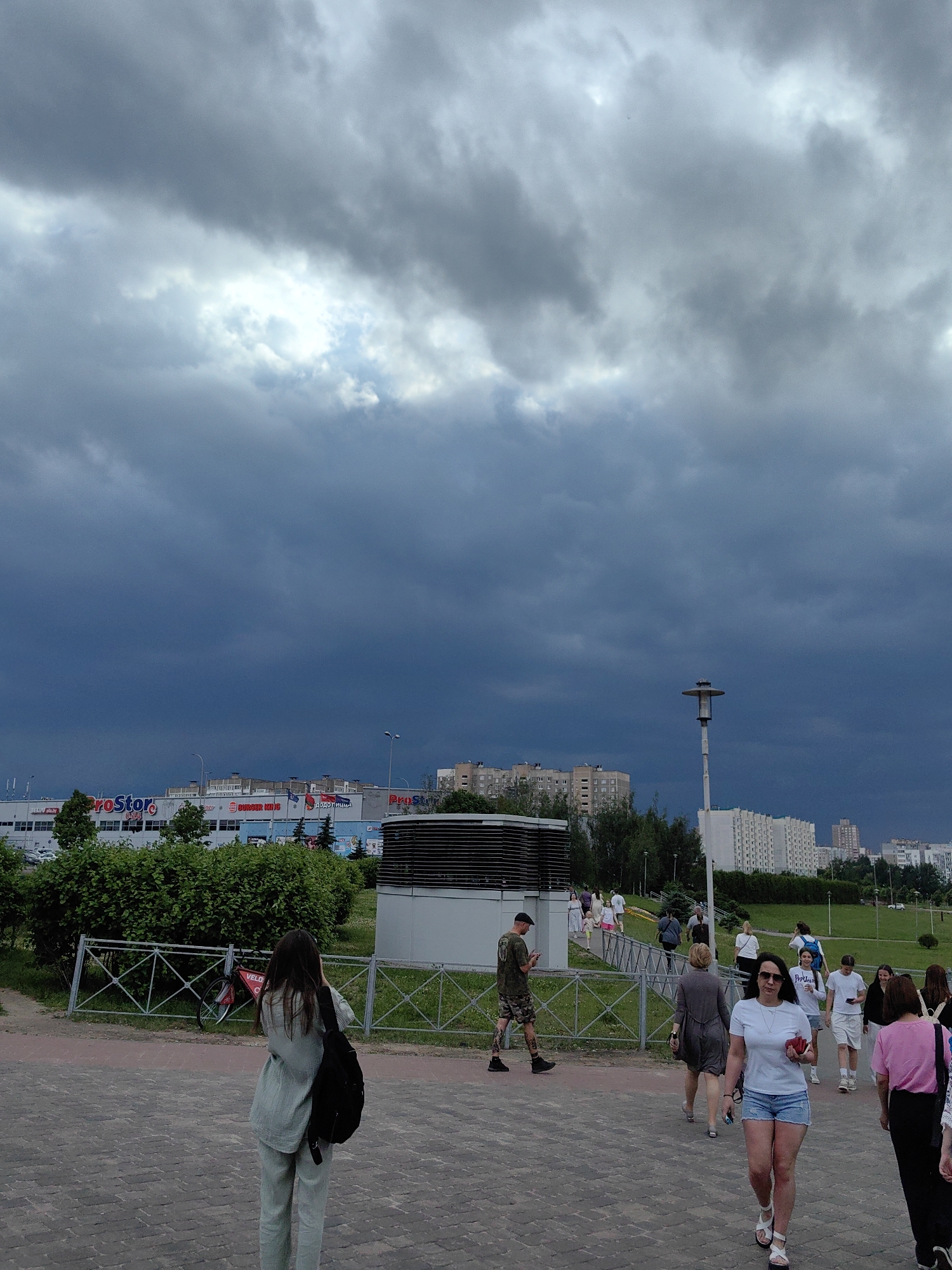 The image depicts a street scene with a large, overcast sky dominating the upper portion. Dark, heavy clouds suggest impending rain. In the middle ground, a commercial building complex is visible with signage for "ProStore" and "Burger King". The building is modern with large windows. Adjacent to the building is a green, grassy area with trees and some outdoor ventilation equipment. A paved walkway is in the foreground, with several individuals walking in various directions. One person in the foreground is wearing a light green outfit and a black backpack, facing away from the camera. Other individuals are dressed in casual summer clothing. A bicycle is leaning against a metal fence. In the background, apartment buildings are visible, characteristic of urban landscapes. The overall impression is a busy urban environment under a dramatic sky. The specific location in Minsk, Belarus can be inferred from the "ProStore" signage which is a well-known supermarket chain in Belarus. This particular ProStore is located on Dzerzhinsky Avenue in Minsk.