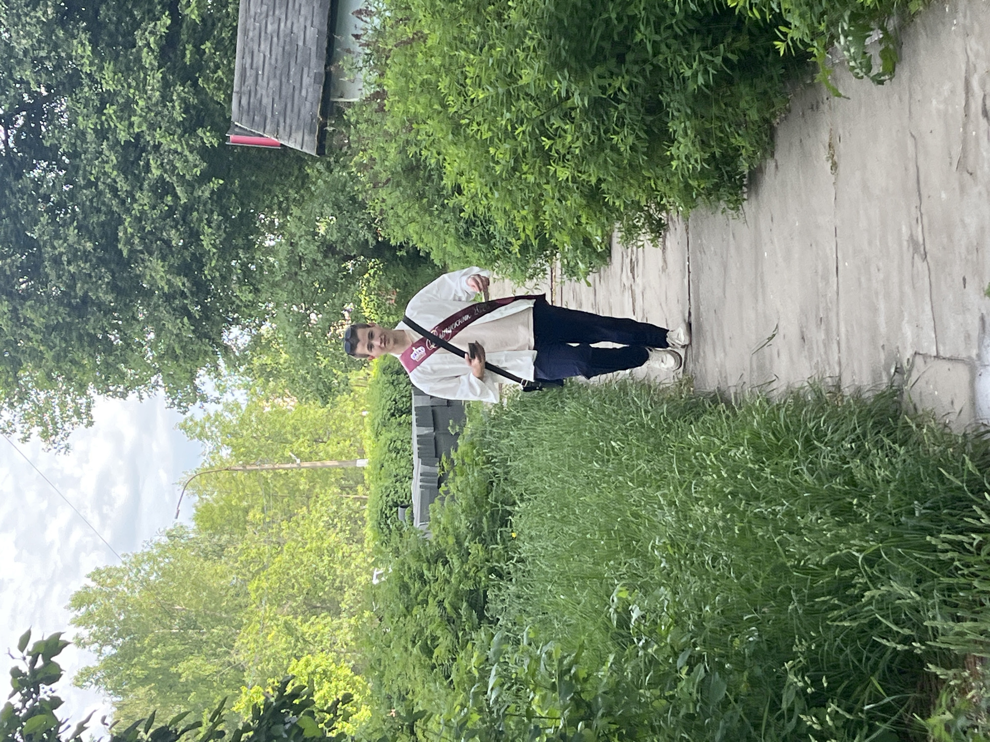A young man stands on a sidewalk in what appears to be a residential area. He is wearing a white shirt, dark pants, and white sneakers. He has a sash across his chest. He carries a long object in his hands, possibly a rod or cane. The environment is overgrown with lush vegetation, with trees and shrubs lining the sidewalk. There is a building visible in the background with a gray roof and red trim. A lamp post is visible. The scene suggests a late spring or summer season. The location is in the city of Chaikovsky, Russia.