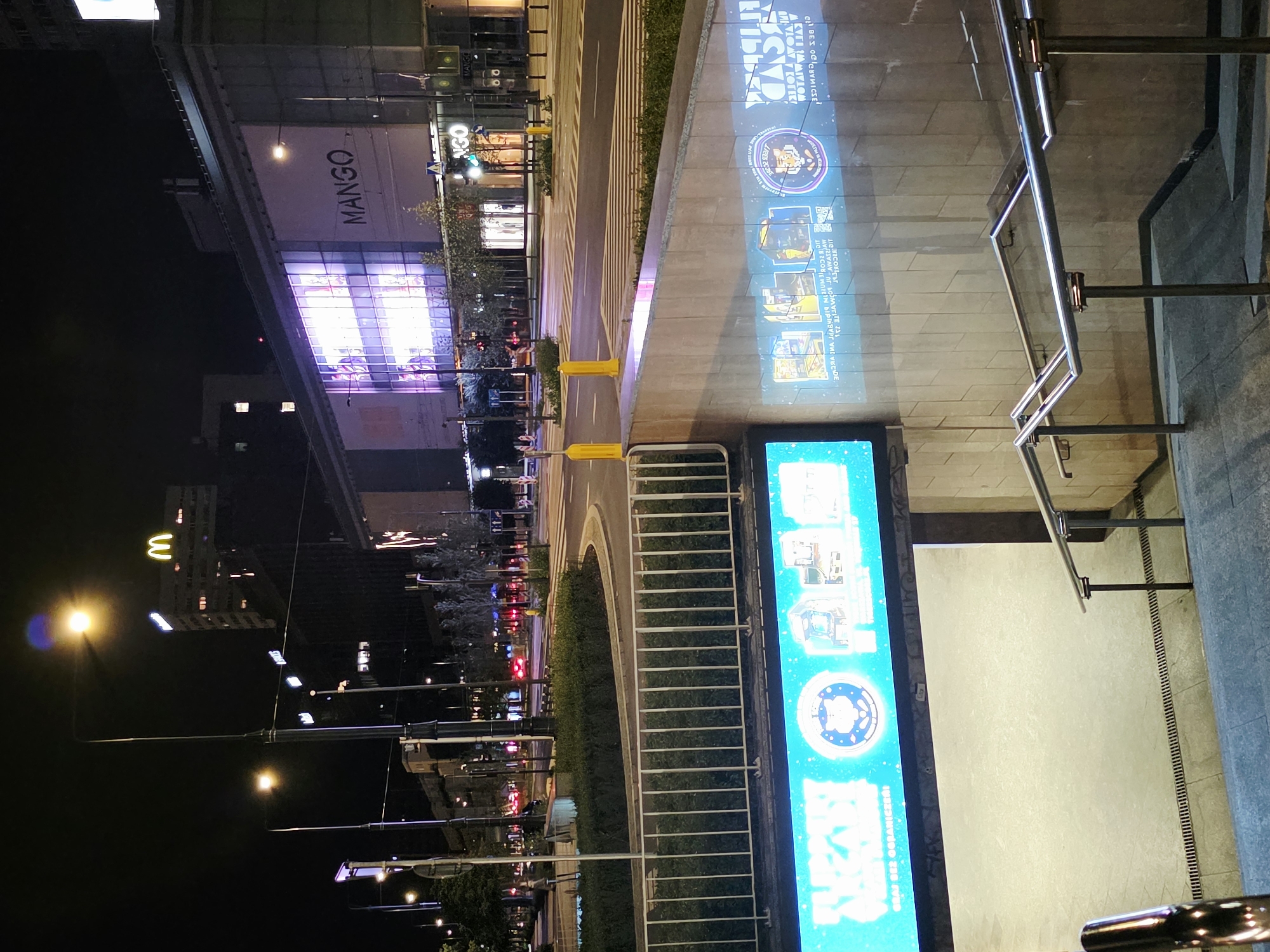 The image shows a nighttime street view in Warsaw, Poland, featuring illuminated buildings and streetlights. Prominently displayed on a large building facade is the "MANGO" logo, suggesting a retail establishment. Below this, a street crosses the scene with traffic lights and pedestrian crossings. In the foreground, a set of stairs leads down to an underground area, highlighted by a large, brightly lit digital screen displaying various graphics and text, including what appear to be images of arcade games and circular emblems. The overall scene depicts an urban environment at night, with a focus on commercial signage and architectural elements.