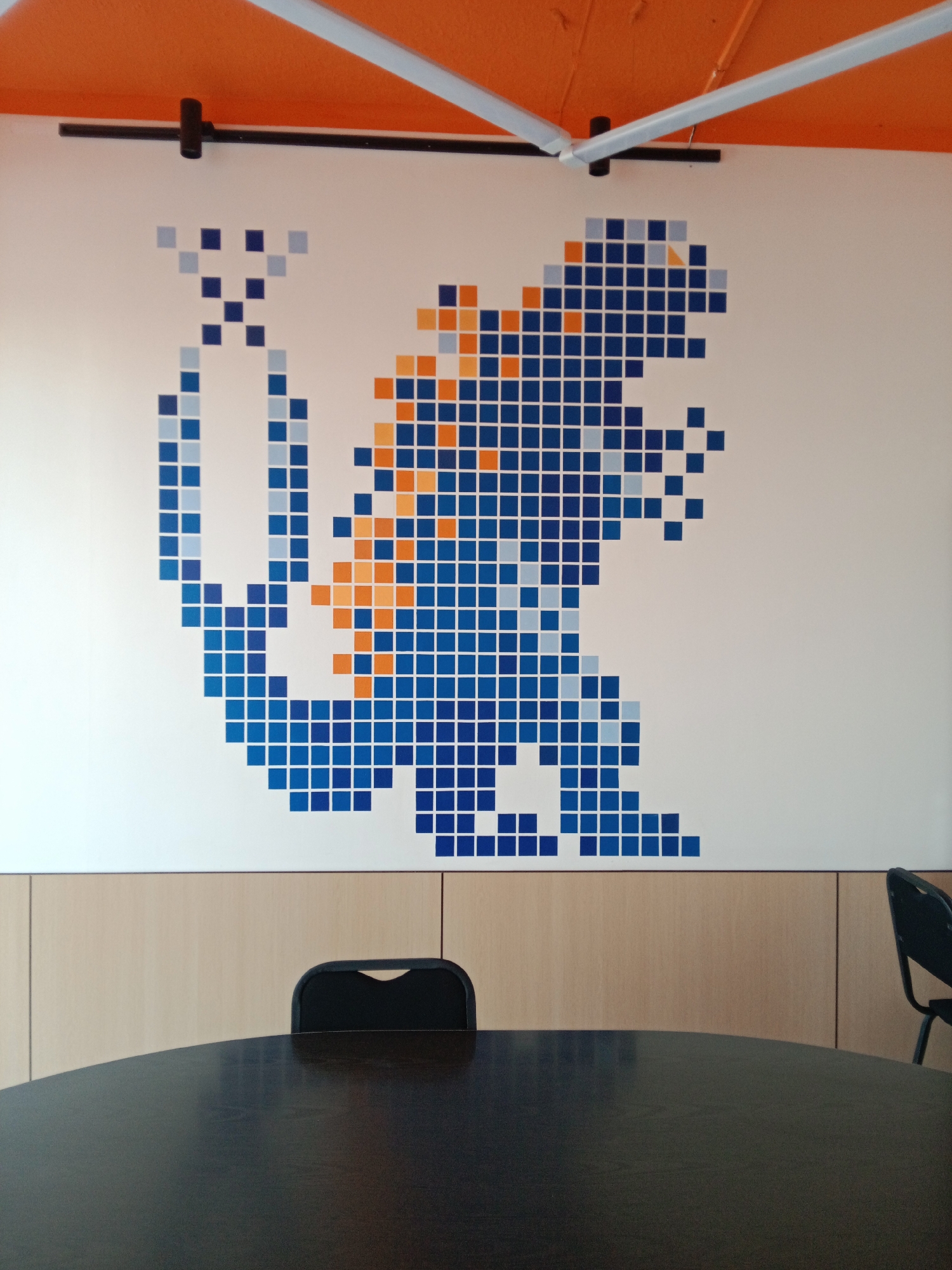 A pixelated mosaic depicting a blue and orange dinosaur is displayed on a white wall. The dinosaur is rendered in shades of blue, with accents of orange and light blue. The mosaic covers a significant portion of the wall. Below the wall is a wooden paneling. In the foreground, a dark, round table is visible, and behind it, a black office chair is positioned. The ceiling is orange. The scene appears to be an indoor setting, possibly an office or meeting room. No specific location in Tashkent, Uzbekistan is discernible from the image.