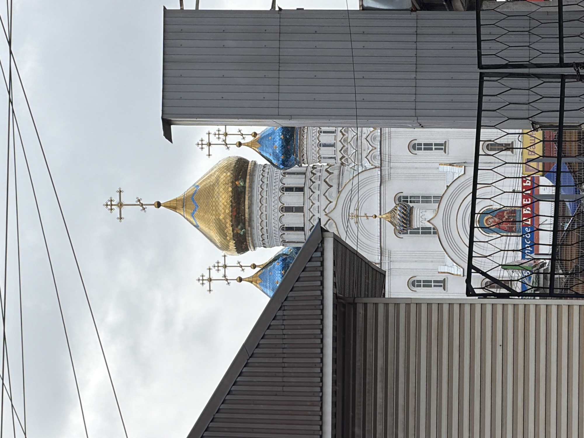 The image captures a view of the Cathedral of the Icon of the Mother of God "Joy of All Who Sorrow" in Liski, Russia, partially obscured by urban elements. The golden and blue domes of the cathedral stand out against the overcast sky. The white walls and decorative details of the building are visible, along with cross-topped spires. The foreground is dominated by the gray, corrugated metal structures of nearby buildings and overhead power lines, which partially block the view of the church. A metal fence is also visible on the right side of the image.