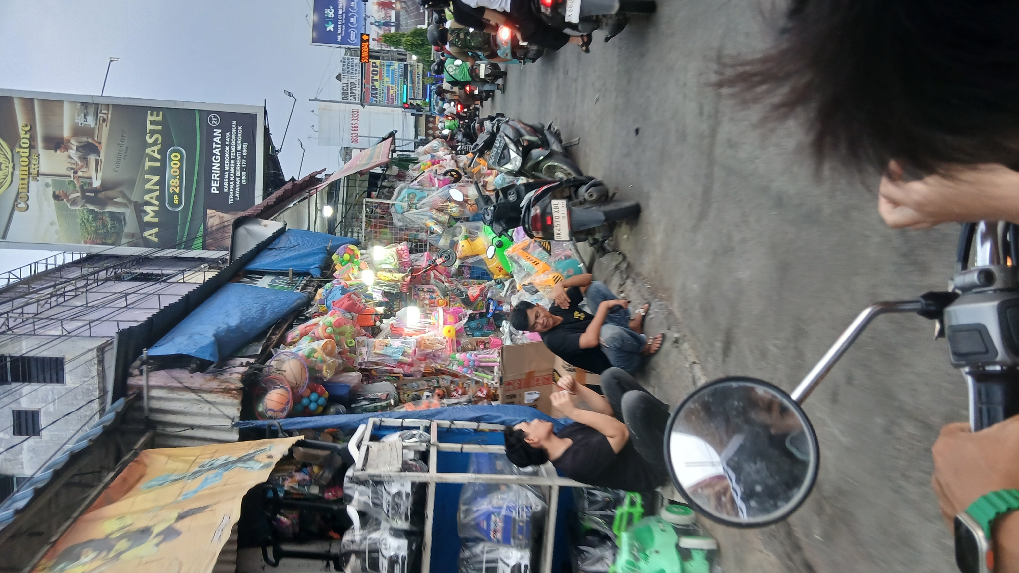 A low-angle, medium shot shows a street scene in the City of Medan, Indonesia, with a focus on a roadside stall filled with colorful toys and a billboard advertisement. Two men are seated on the ground in front of the stall. The man on the left, wearing a black t-shirt and gray pants, gestures with his hands as he speaks. The man on the right, also in a black t-shirt and blue jeans, leans forward with his right arm resting on his knee. A motorcycle with the license plate "DK 2570 XBL" is parked to their right. Behind the stall, a large billboard for "Commodore Filter" is prominently displayed, featuring an advertisement for "A MAN TASTE" priced at RP 28.000, and a warning about smoking. The street is busy with motorcycles and other vehicles, indicating a lively urban environment. The lighting suggests it is late afternoon or early evening.