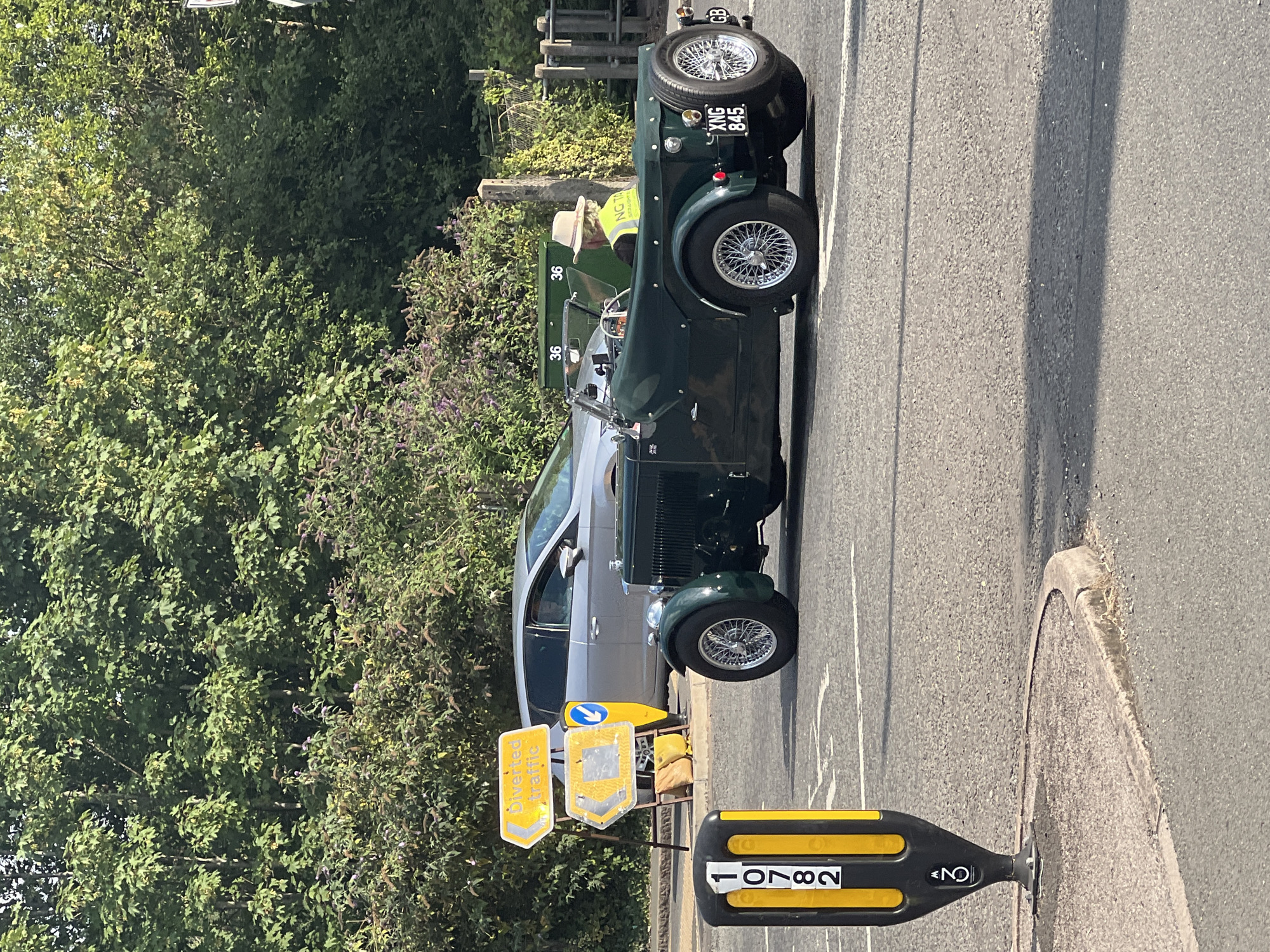 A green vintage convertible car is parked at an intersection in Guildford, United Kingdom. The car, with license plate XNG 845, has a spare tire mounted on the rear. The driver, wearing a yellow high-visibility vest and a straw hat, is seated in the car. In the background, a silver hatchback is parked, and traffic signs indicate a diversion. A black and yellow bollard with the number 10782 is visible in the foreground. The scene is illuminated by sunlight, casting shadows on the asphalt road.