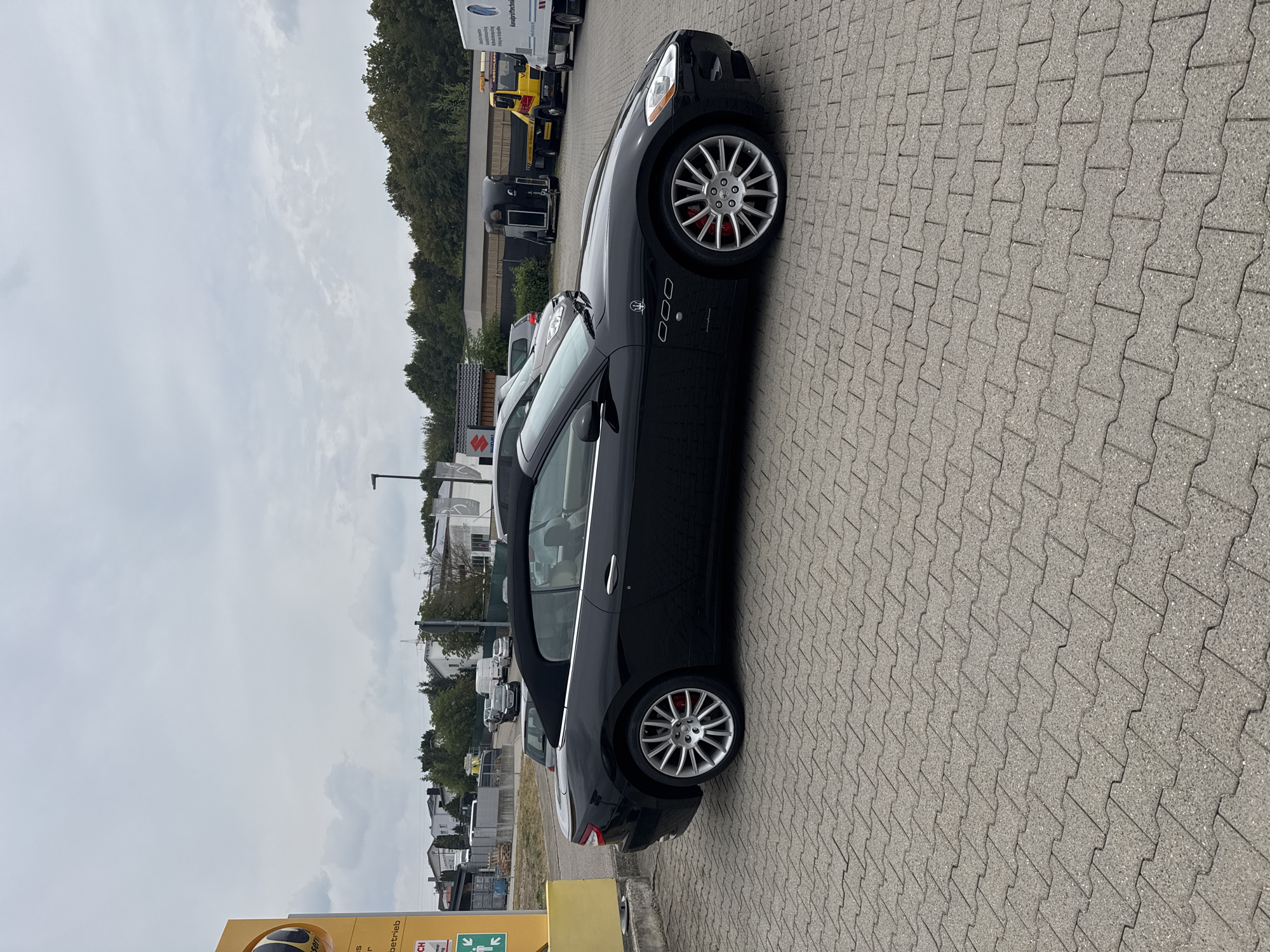 A black convertible Maserati GranTurismo is parked on a grey paver stone lot in front of a building with a yellow sign that reads "Autohaus". The car is facing right and is viewed from the side. The convertible top is up. The car has silver multi-spoke wheels with red brake calipers. In the background, there are other vehicles, including a yellow tow truck, a small black trailer, and a white truck. The sky is overcast. The location is Wettstetten, Germany.