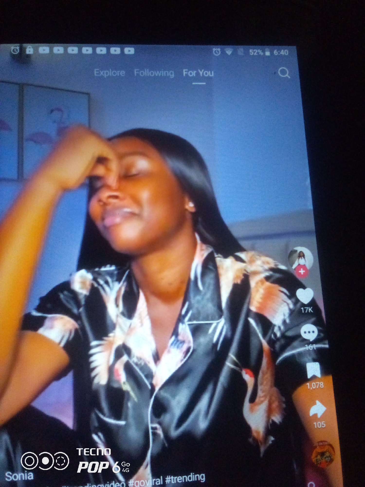 The image shows a close-up of a woman wearing silk pajamas with a crane pattern. She is resting her hand on her forehead and her eyes are closed. The background is a blue wall with two framed pictures of flamingos. On the right side of the screen, there are social media icons for likes, comments, shares, and a user profile. At the bottom left, there is a Tecno Pop 6 4G logo and the text "Sonia". The top of the screen displays the time as 6:40, a battery icon indicating 52% charge, and navigation tabs for "Explore", "Following", and "For You". The scene appears to be indoors, likely in a bedroom. The image is a screenshot from a mobile device.