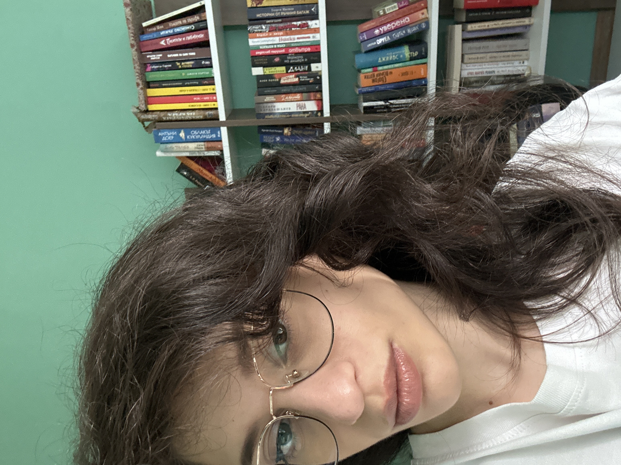 The image is a close-up, over-the-shoulder selfie taken indoors. The foreground is dominated by the left side of a young woman's face and her dark, wavy hair, which obscures part of the background. She is wearing round, gold-rimmed glasses and a white t-shirt. Her lips are slightly parted, and her gaze is directed towards the camera.
Behind her, a portion of a white bookshelf is visible, filled with books of various sizes and colors. The books are arranged tightly on multiple shelves. The wall behind the bookshelf is painted a vibrant teal color. The lighting suggests an indoor environment, possibly a home or study. The scene provides no specific indicators for a location in Sofia, Bulgaria.