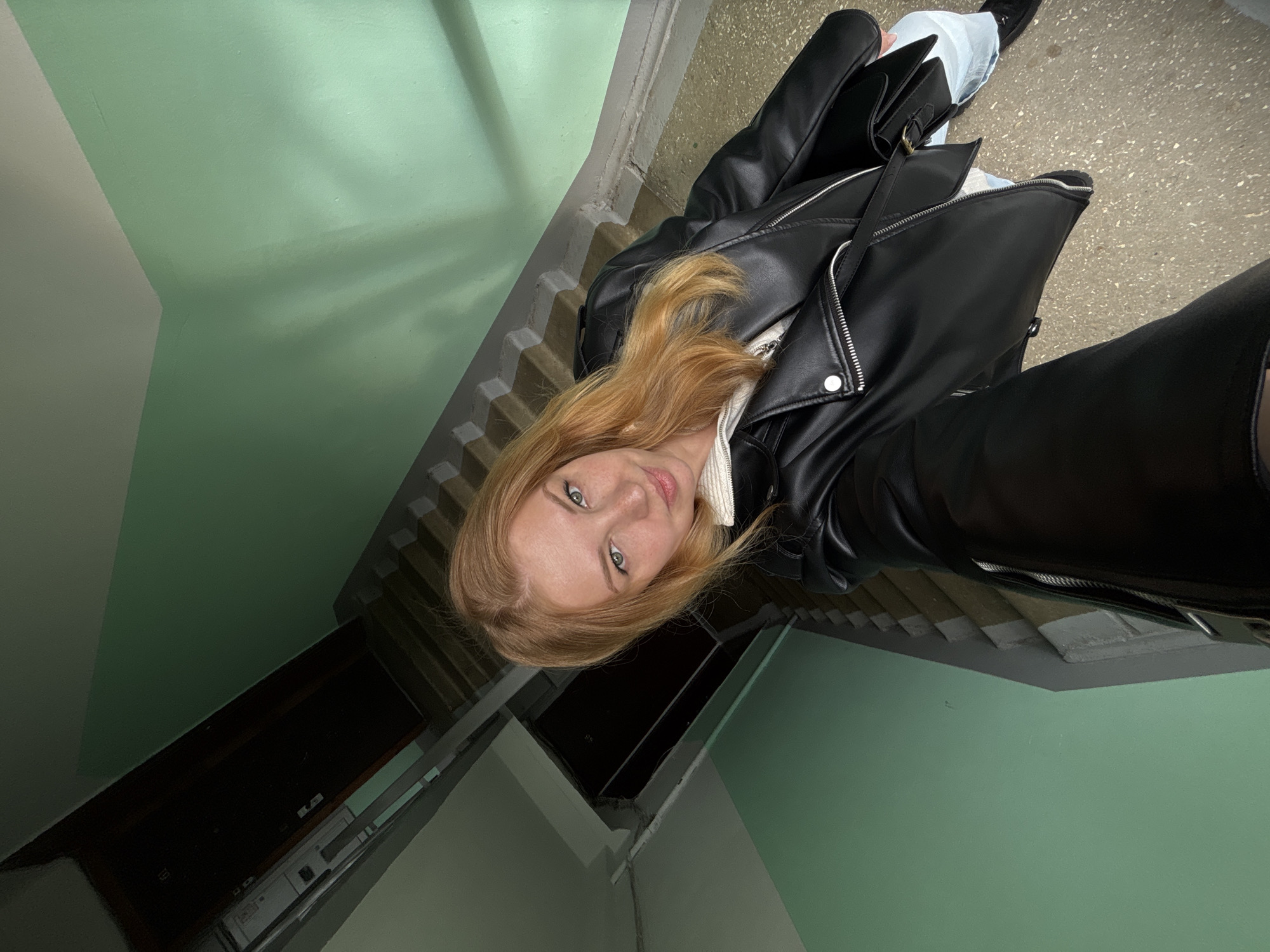 A person with strawberry blonde hair, wearing a black leather jacket and light blue jeans, stands on a gray stairwell with green walls. The photograph is taken from a high angle, looking down at the subject. The location is unknown.