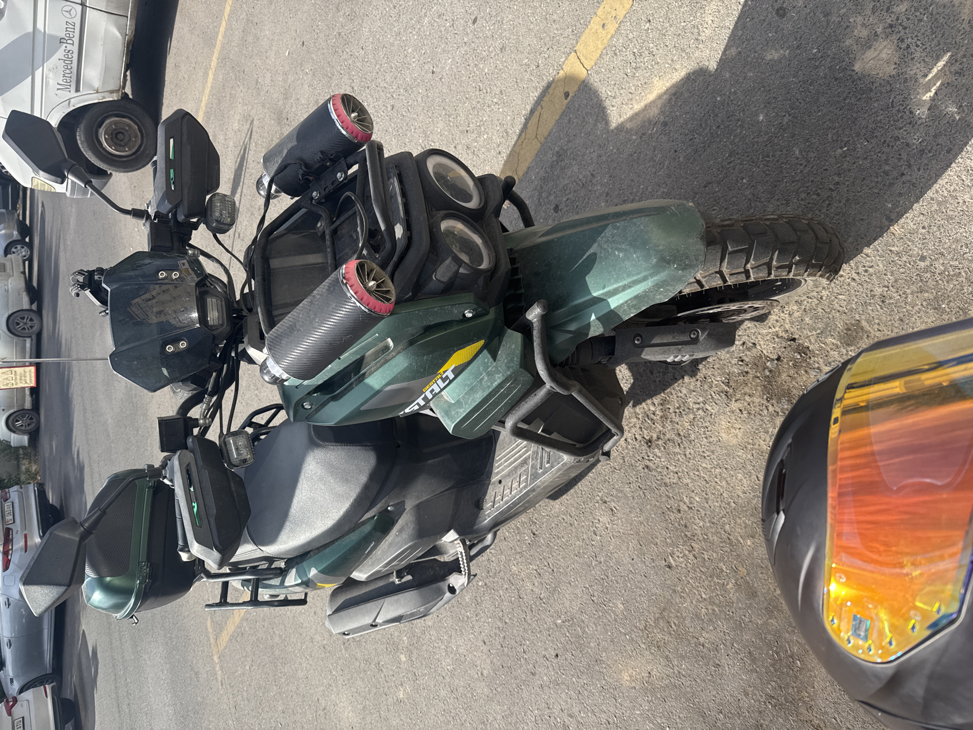 An eye-level medium shot captures a dark green scooter parked on an asphalt surface marked with yellow lines, likely in a parking lot. The scooter is equipped with a large black rear storage box, black handguards with integrated green lights, and dual round headlights. Two cylindrical, carbon fiber-like accessories with red accents are attached to the handlebars. A black motorcycle helmet with a bright orange-tinted visor is positioned in the foreground, partially obscuring the scooter's front wheel. In the background, several cars are visible, including a white Mercedes-Benz Sprinter van. The scene is well-lit, indicating it is daytime, and the sun casts distinct shadows.