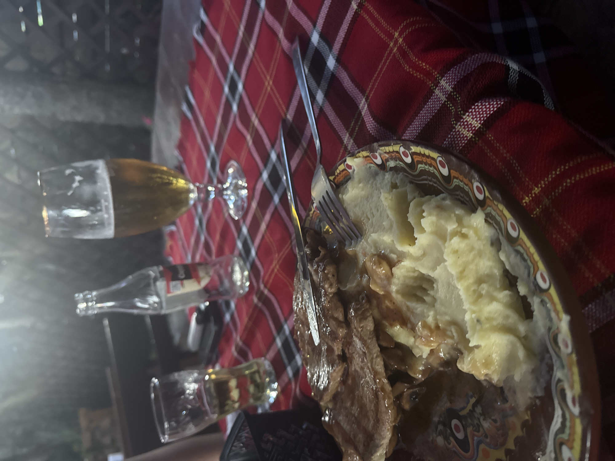 The image depicts a meal at an outdoor restaurant in Sofia, Bulgaria. The table is covered with a red, black, and white plaid tablecloth. In the foreground, a plate of food is visible, containing several slices of meat topped with sauce and mushrooms, accompanied by mashed potatoes. A knife and fork are placed on the plate. To the left, a glass with a light-colored beverage, likely beer, sits next to a clear glass bottle. To the right, another glass, also containing a light-colored beverage, is partially visible. In the background, out-of-focus elements suggest a rustic outdoor setting with a dark, textured wall or fencing. The lighting is dim, with some light reflecting on the glasses and the food. There are no people clearly visible in the frame.