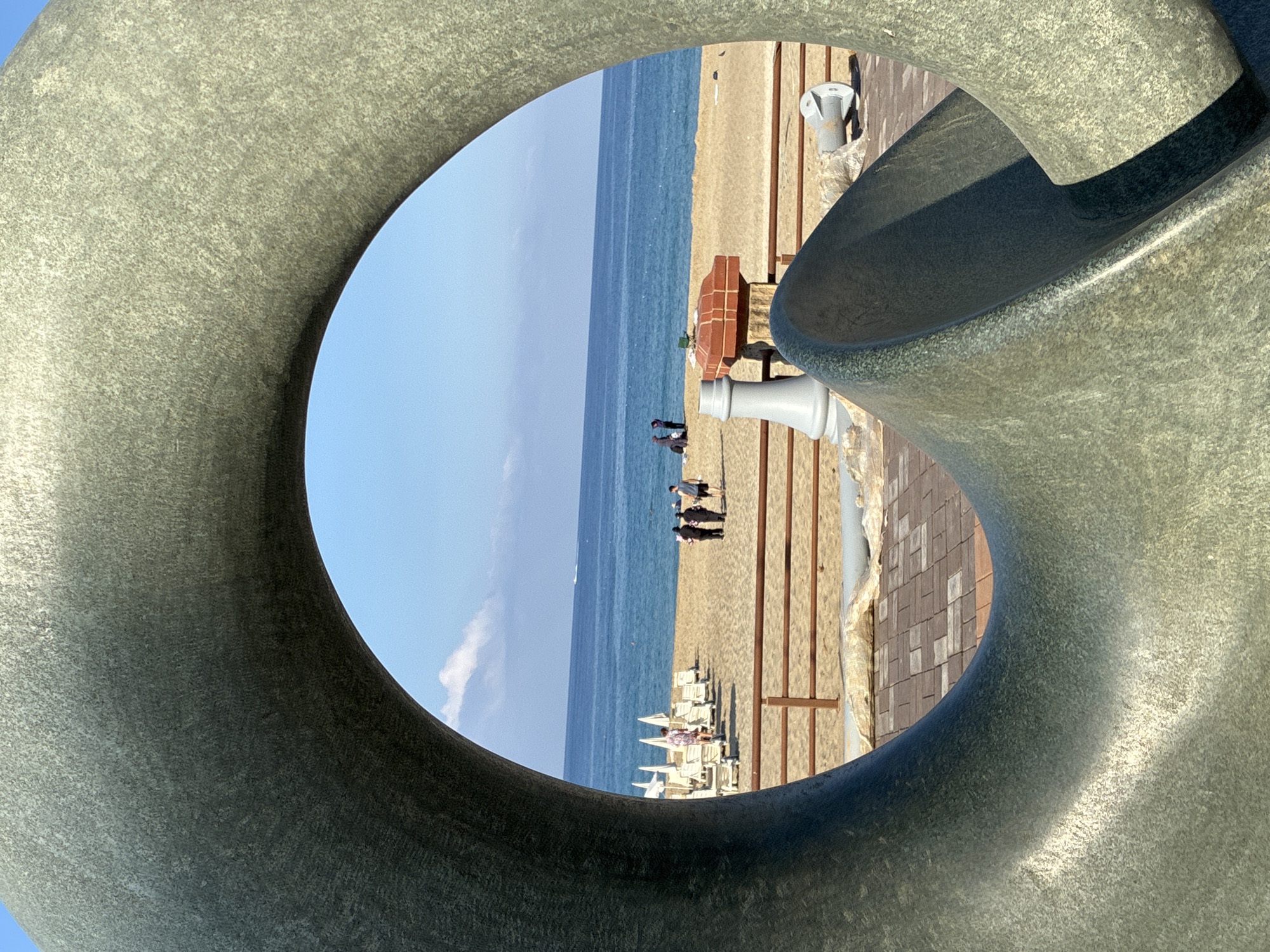 The image captures a view of the beach and the sea through a large, grey, circular metal sculpture. The sculpture is textured and appears to be made of stone or metal. In the background, the sandy beach is lined with rows of empty lounge chairs and umbrellas, suggesting a beach resort. Several people are visible on the beach, walking and standing. The blue sea stretches to the horizon under a clear blue sky with a few scattered white clouds. The foreground features a paved walkway with some architectural elements and a wooden railing separating it from the beach. This scene is likely located in Alanya, Turkey, given the typical coastal scenery and potential for public art installations in tourist areas.
