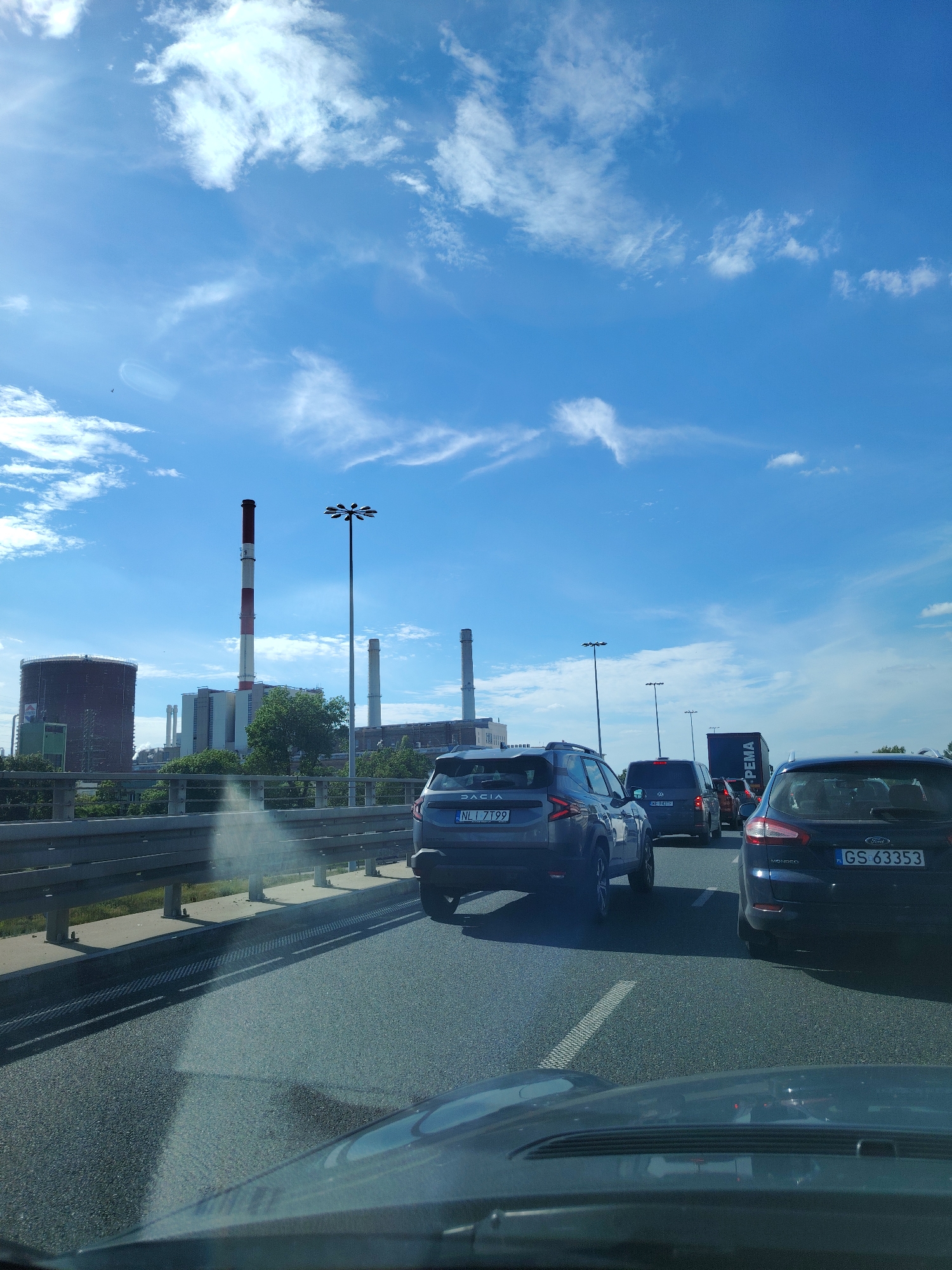 The image depicts a traffic jam on an elevated road in Warsaw, Poland, with a power plant in the background. The sky is blue with scattered white clouds. The power plant features several tall, striped smokestacks and a large, cylindrical storage tank. Several cars are visible on the road, including a dark blue Ford Mondeo with license plate GS 63353 and a grey Dacia with license plate NL1 7T99. A large, dark truck with "PEMA" written on its side is also present. The road is paved, and a metal guardrail separates the lanes. The perspective suggests the photo was taken from the driver's seat of a vehicle.