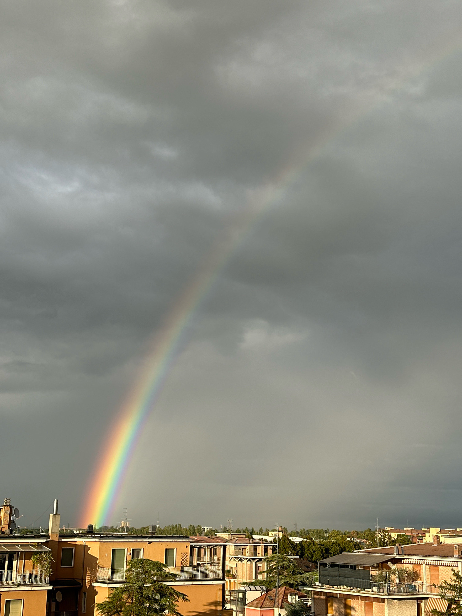 An overcast sky with a vibrant rainbow stretches across the frame, its arc beginning in the lower left quadrant and extending upwards and to the right. Below the sky, a cityscape of buildings with orange and terracotta roofs is visible, interspersed with trees. The buildings appear to be residential structures with balconies and windows. The scene captures a moment after a rain shower, with the sun breaking through the clouds to create the rainbow. The overall impression is one of a dramatic natural phenomenon over an urban environment in Parma, Italy.
