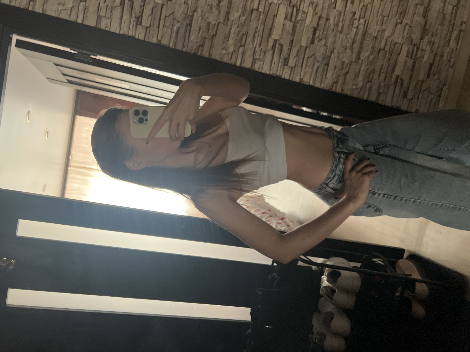 A young woman is taking a mirror selfie. She is wearing a white top and jeans. The background appears to be a room with a stone wall. A shoe rack filled with shoes is visible in the lower part of the frame. The location is indiscernible from the information given. Noviy, Russia cannot be determined.