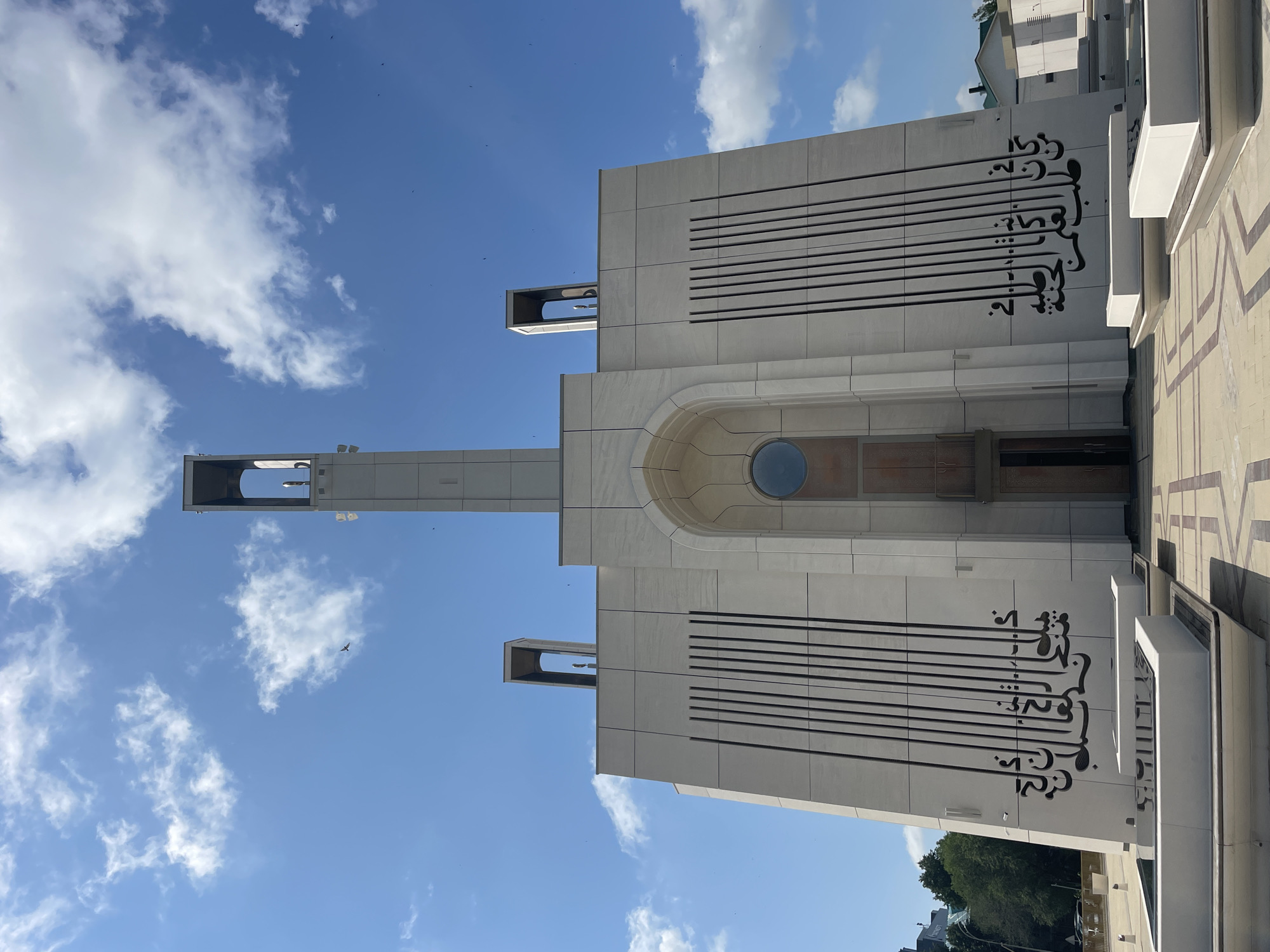 The image shows a modern, white mosque with a tall, slender minaret. The mosque features geometric patterns and Arabic calligraphy on its facade. In front of the mosque, there are low, white structures that appear to be fountains or seating areas. The ground is paved with a patterned tile. The sky is blue with scattered white clouds, and a bird is visible flying in the distance. The mosque is located in Almetyevsk, Russia, as indicated by the context.