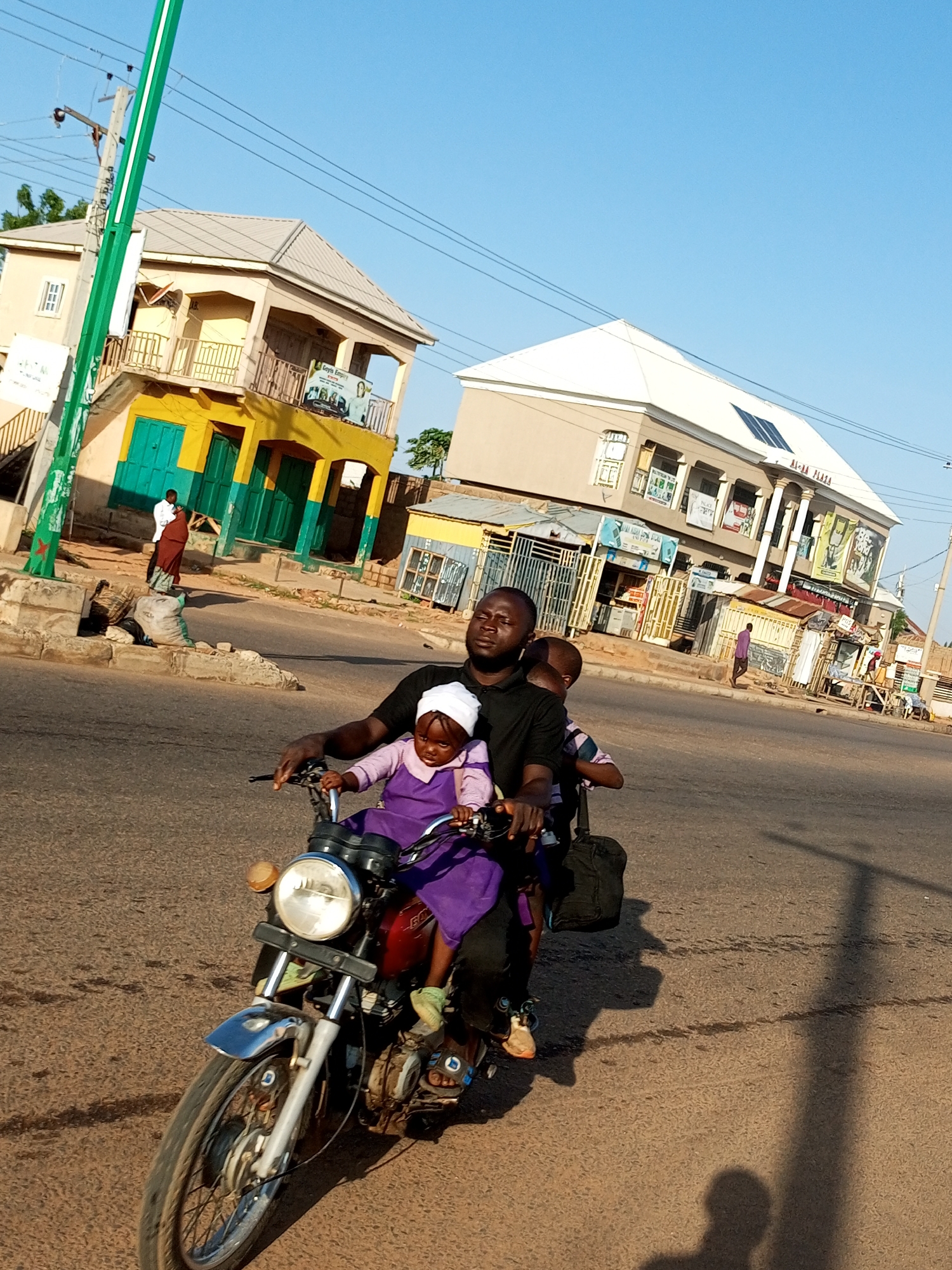 An eye-level, medium shot captures a man driving a motorcycle with three children as passengers on a paved road in Garko, Nigeria. The driver, a dark-skinned man wearing a black t-shirt, is in the foreground on the left, looking to the right. A young child wearing a white head covering and a purple dress is seated in front of him, also facing right. Behind the driver, another child, partially obscured, sits on the motorcycle. The motorcycle is dark red with a chrome fender and a round headlight. In the background, there are buildings with yellow and cream facades, some with balconies and posters displayed. A green pole with white stripes is visible on the left side of the frame. The sky is clear and blue.