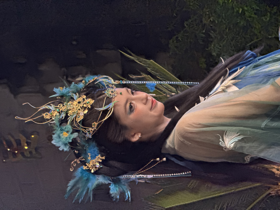 The image depicts a young adult female, of East Asian descent, posing in elaborate traditional attire within the Yuzhong District, China. She is shown in a three-quarter profile, facing towards the lower right of the frame, with a soft, slightly smiling expression. Her dark hair is styled in an ornate updo, featuring an elaborate golden headpiece adorned with numerous teal and blue feathers, intricate gold filigree, and small green and clear gemstones. Multiple long, shimmering blue beaded chains and shorter gold chains with small charms hang from the headpiece. Her makeup includes blue eyeshadow, subtle glitter beneath the eyes, and a small red accent above the outer corner of each eye. She wears layered traditional Chinese garments; the visible outer layer is a sheer, light teal-green fabric with delicate white or silver embroidery depicting a bird-like motif on the left shoulder/sleeve. A blue and teal inner garment is visible beneath. The background is dimly lit and dark, with blurred green foliage, possibly large leaves or ferns, visible behind and to the right of the subject. In the upper left background, a faint, blurred warm light source, potentially an illuminated architectural detail or sign, is discernible. The individual is stationary, posing for a photograph, and no other persons or direct interactions are visible.