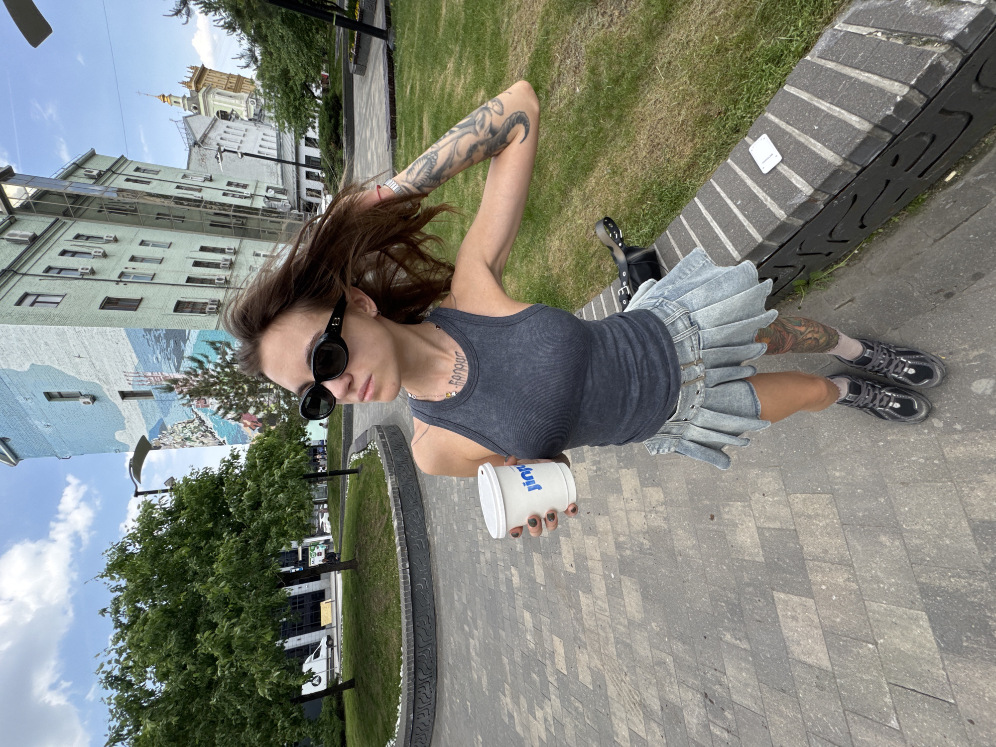 A young woman stands in a park-like setting with trees and buildings in the background. The woman is wearing a gray tank top, a denim mini skirt with pleats, black sneakers and socks, and dark sunglasses. She has tattoos on her arm, neck, and leg. She is holding a white cup with the word "Joy" printed on it. The cup appears to be in her right hand. The woman appears to be standing on a gray paved walkway. To the right of the woman is a gray bench. The background shows buildings, some with light-colored facades and many windows. The scene appears to be Moscow, Russia, based on the building architecture, a historic bell tower structure, and the typical urban park setting in Moscow.