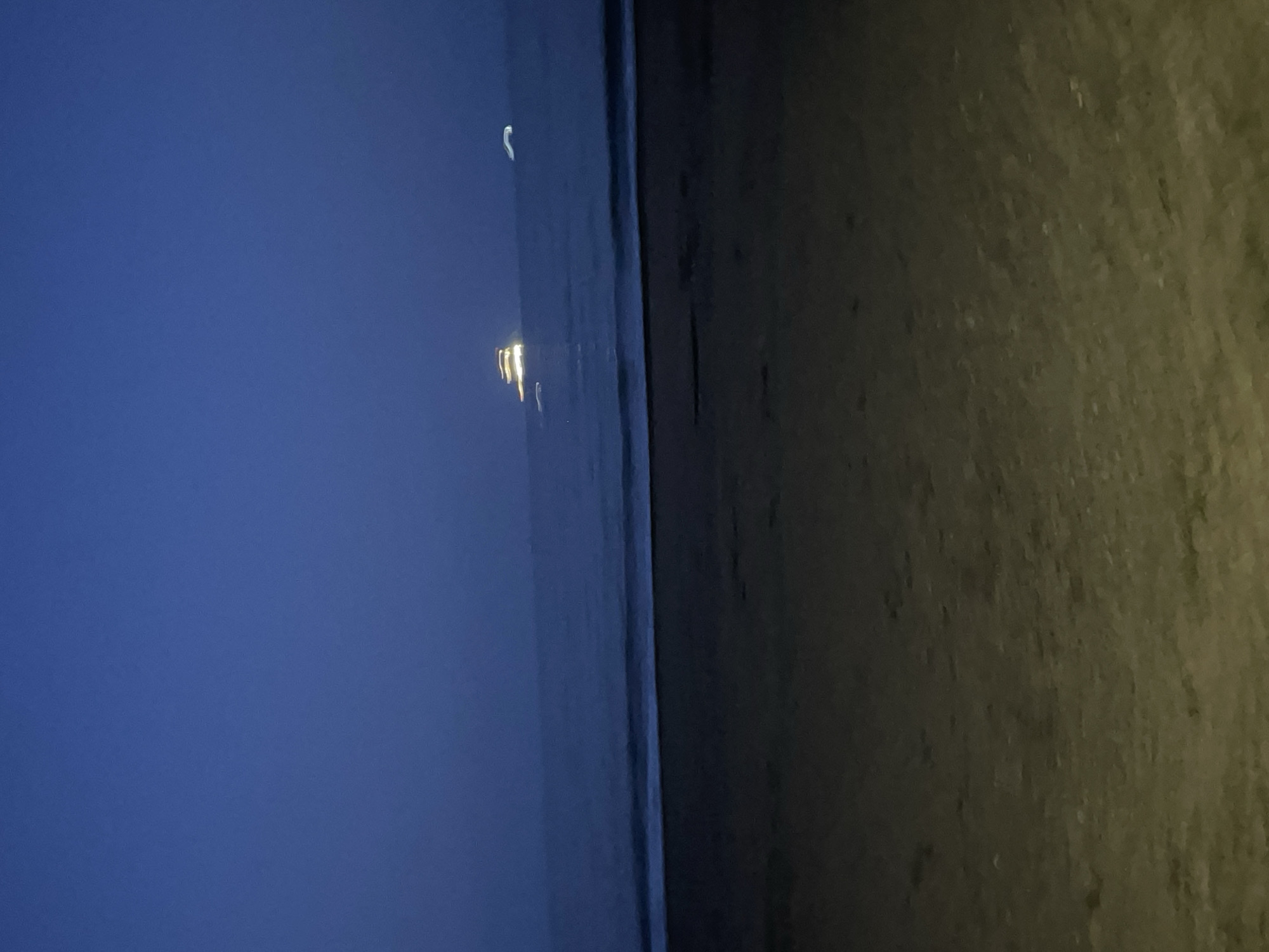 The image shows a beach at night, likely in Burgas, Bulgaria. To the left is a dark blue sky, possibly with a partial moon visible. In the middle is the dark sea, with blurred lights on the horizon, potentially from a ship or distant buildings. To the right is the beach itself, appearing grainy and dark, with textures suggesting sand. There are no discernible people or actions visible. The overall environment is dimly lit and suggests a night scene by the sea.