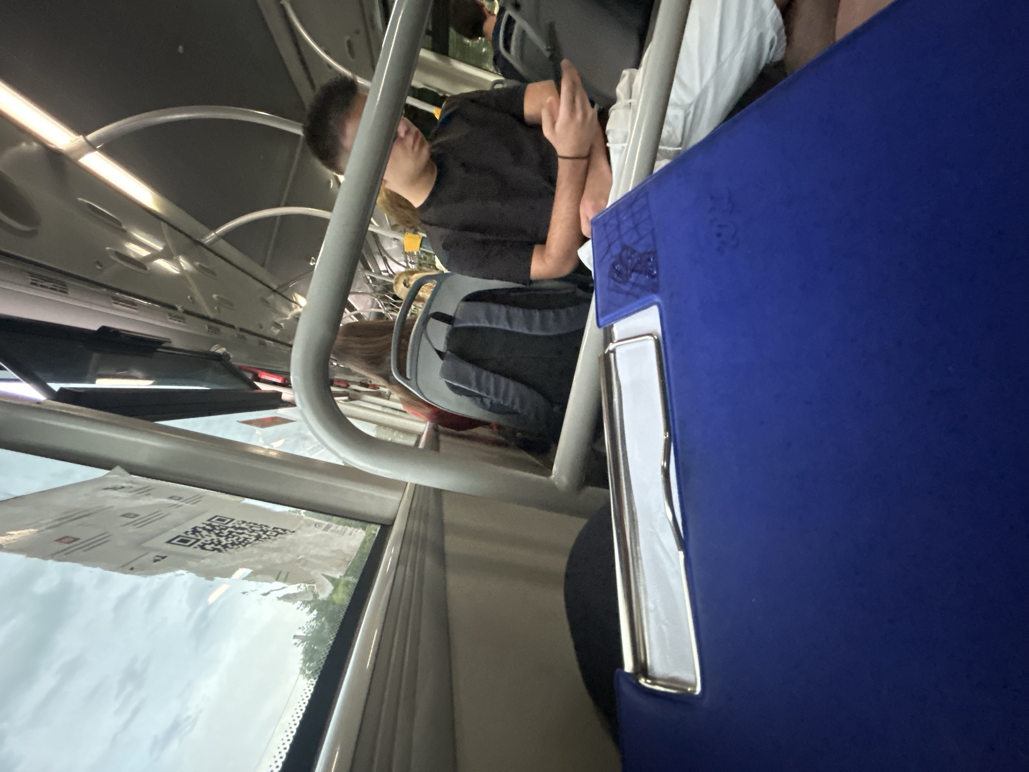 The image shows the interior of a bus. In the foreground, a blue binder with a silver clasp is positioned on a seat. The binder has a faint embossed logo. A young man wearing glasses and a black t-shirt is seated in the middle ground, looking down at a black smartphone he is holding in his hands. He is wearing white pants. Behind him, a gray backpack is visible on a seat. Further back, other passengers are seated, but their faces are not clearly visible. The bus has overhead handrails and fluorescent lighting. Through the windows, the sky appears cloudy, and some trees and buildings are visible outside, suggesting an urban or suburban environment. A QR code sticker is attached to the window on the left side of the image.