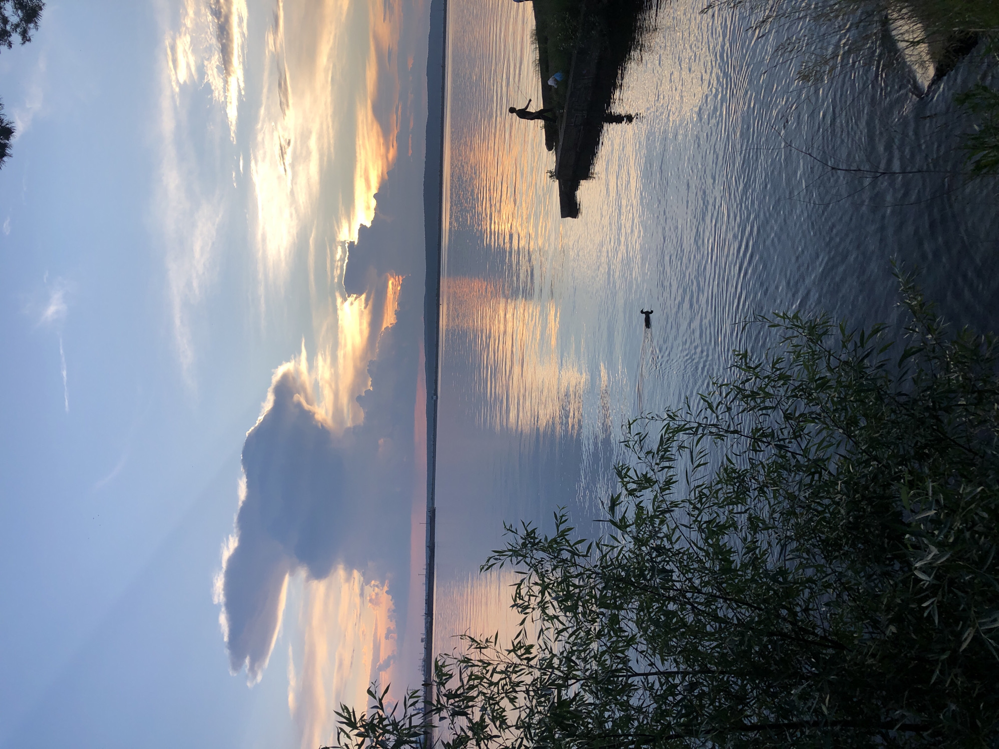 The image depicts a serene sunset over a wide body of water, likely the Kama River, in Chaikovsky, Russia. The sky is painted with soft hues of orange, pink, and blue, with dramatic cloud formations casting shadows and catching the last rays of sunlight. On a concrete embankment extending into the water, a lone figure is silhouetted against the vibrant sky, appearing to be fishing with a rod. To the right of the fisherman, a small child is sitting on the embankment. In the foreground, lush green foliage partially obscures the view of the water, with a duck swimming across the calm surface. In the distance, a hazy shoreline with industrial structures can be observed, suggesting the urban context of Chaikovsky. The overall atmosphere is tranquil and picturesque.