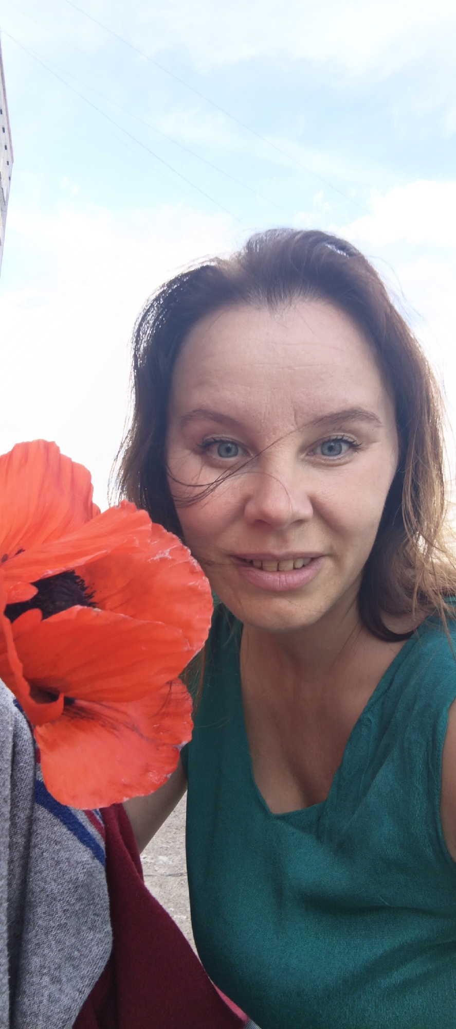 A woman with brown hair and blue eyes smiles at the camera, holding a large red poppy in her left hand. She is wearing a teal-colored dress. A gray and maroon blanket is visible draped over her left arm. The background is a bright blue sky with some clouds, and a portion of a building is visible on the far left. The photo was taken outdoors on a bright day.