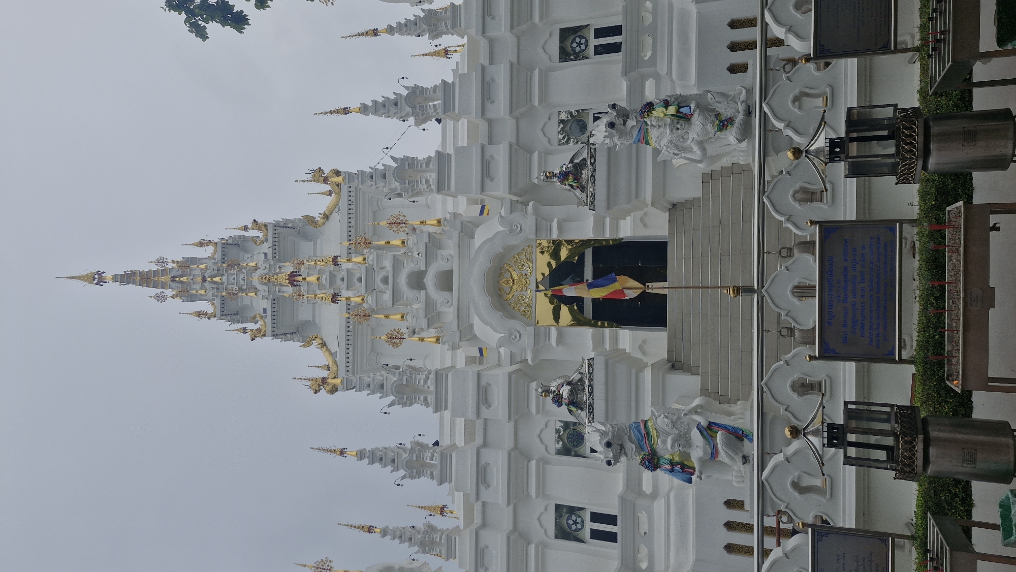A full-shot, eye-level, vertical photograph captures the ornate white facade of a Thai temple, Wat Phra That Doi Suthep, situated in the mountainous region of Ban Pong Nuea, Thailand. The temple's architecture is characterized by multiple tiered roofs, adorned with golden spires and intricate detailing. A central staircase leads to the main entrance, flanked by two guardian statues that appear to be mythical beasts. A Thai flag stands prominently in front of the entrance, which features a golden-colored door. Decorative elements, such as statues of deities and elaborate moldings, are visible throughout the structure. In the foreground, a row of incense burners and a metal lantern contribute to the spiritual ambiance. The sky above is overcast, and a few branches of a tree are visible on the right side of the frame, suggesting an outdoor setting. The overall scene conveys a sense of grandeur and religious devotion.