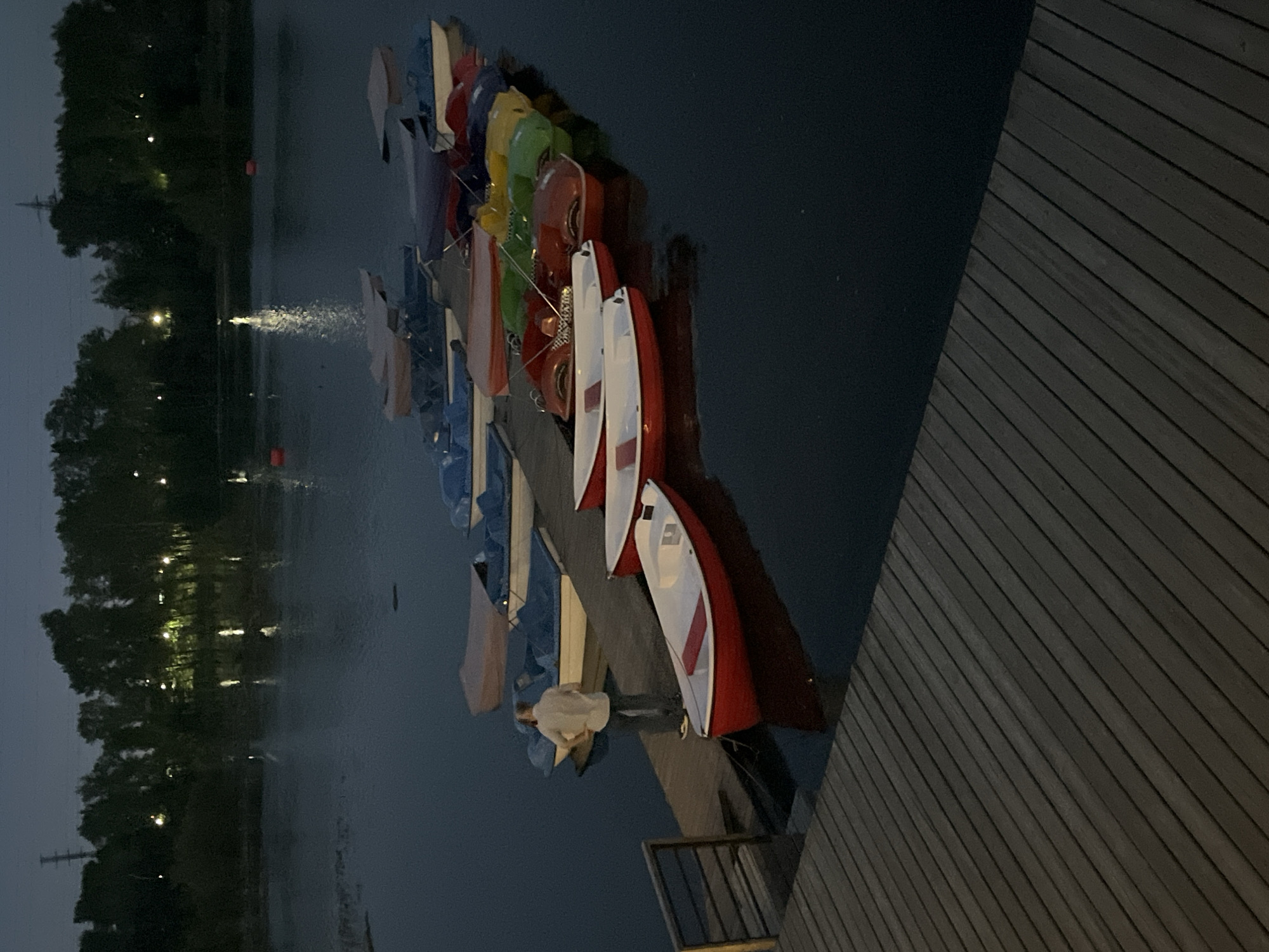 A group of pedal boats are moored to a wooden pier on a calm body of water at dusk. The water reflects the lights from the trees lining the opposite bank, creating a serene atmosphere. In the foreground, a man in a light-colored shirt and dark pants stands on the pier, facing away from the camera, appearing to be overseeing the boats. The boats are of various bright colors, including red, yellow, green, and blue, and some are covered with tarps. The pier extends into the water, and the wooden planks are visible in the foreground, showing wear and tear. The overall scene suggests a recreational area, possibly a park or a lake in the city of Krasnoyarsk, Russia, where such activities might be popular.