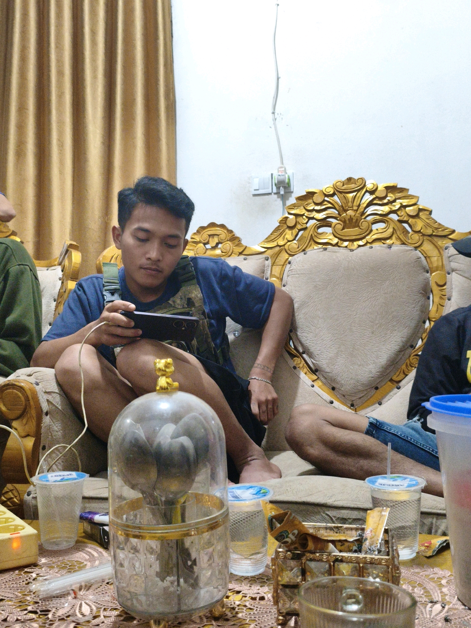 In a dimly lit room, possibly a living room in Koto Baru Semurup, Indonesia, three individuals are seated on a plush, ornate sofa with a golden, carved headrest. The sofa's fabric appears to be a light beige with a subtle patterned texture.
A young man in the center, wearing a dark blue t-shirt and a tactical vest, is engrossed in playing a mobile game on his phone, which is connected to a charger. His legs are crossed, and he is leaning back on the sofa. To his left, another person, partially visible, is wearing a dark green long-sleeved garment. To the right of the central figure, another individual, also partially visible, is wearing denim jeans and a black shirt.
In the foreground, a table is covered with a lace-like tablecloth. On the table, there are several items including plastic cups with clear liquid, a golden decorative item resembling a cat sitting on a clear dome containing spoons, a cigarette pack and lighter, an electrical power strip with an illuminated socket, and a clear glass containing a beverage. The overall ambiance suggests a casual gathering or downtime.