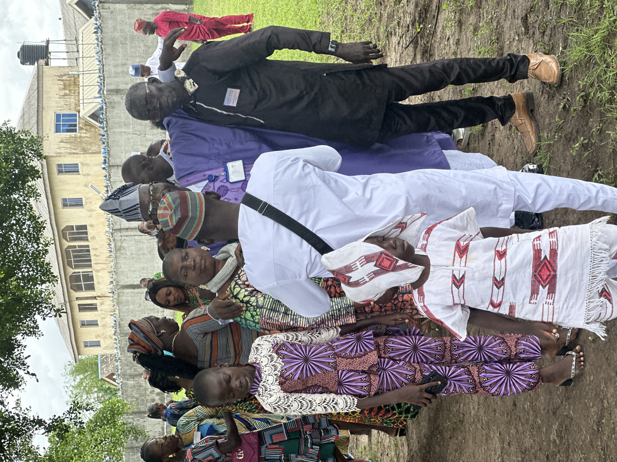 In Itoga, Nigeria, a group of people are gathered outdoors in front of a fenced building. The building has a light yellow facade and multiple windows. A chain-link fence with a concrete base runs along the perimeter. Several individuals are dressed in traditional African attire, featuring vibrant patterns and head coverings. In the foreground, a child in a white and red patterned outfit stands next to a woman wearing a purple and white patterned dress. Behind them, a man in a black tunic and pants, and another in a white tunic, are visible. Several other individuals are seen in the background, some wearing bright clothing and headwraps. The ground is covered with dirt and sparse green grass. The sky is overcast.