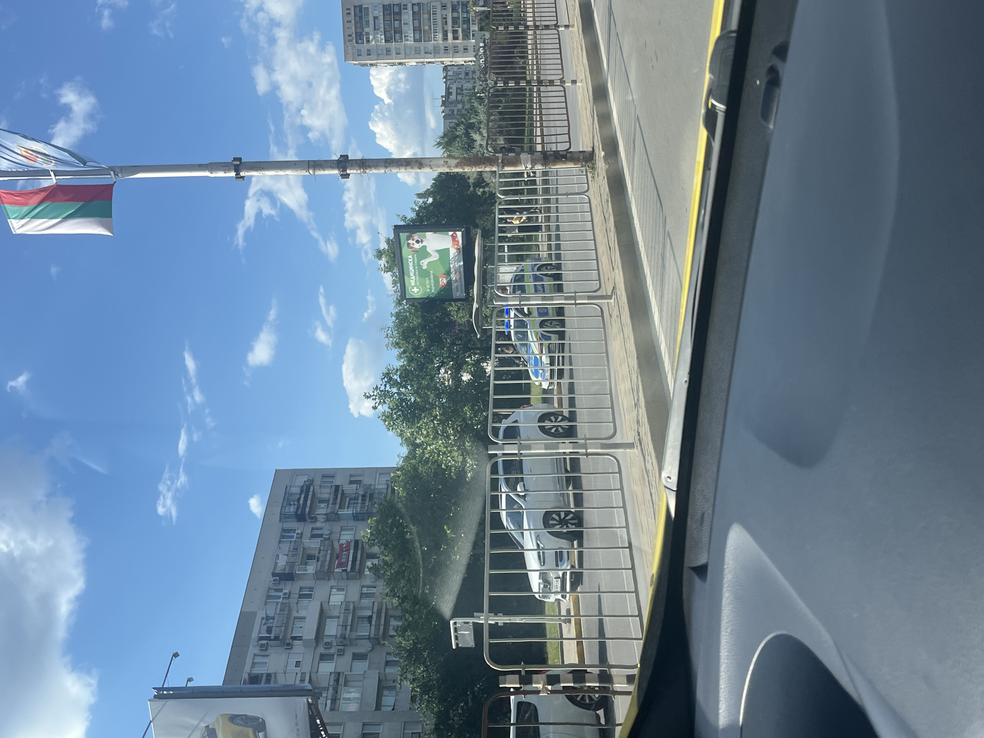 The image shows a street scene in Sofia, Bulgaria, on a sunny day with clouds in the sky. In the foreground, a section of a car's dashboard is visible. Beyond the dashboard, a yellow line is painted on the road, and a metal fence separates the road from a sidewalk.
On the sidewalk, several cars are parked or driving. A white SUV is prominent, and behind it, a police car with flashing blue lights is visible. Further down the street, a billboard displays an advertisement with a dog. Buildings line the opposite side of the street, appearing to be apartment blocks. A flagpole with the Bulgarian flag and another flag is also present. Trees and greenery are scattered throughout the scene. The overall impression is of a typical urban environment in Sofia.