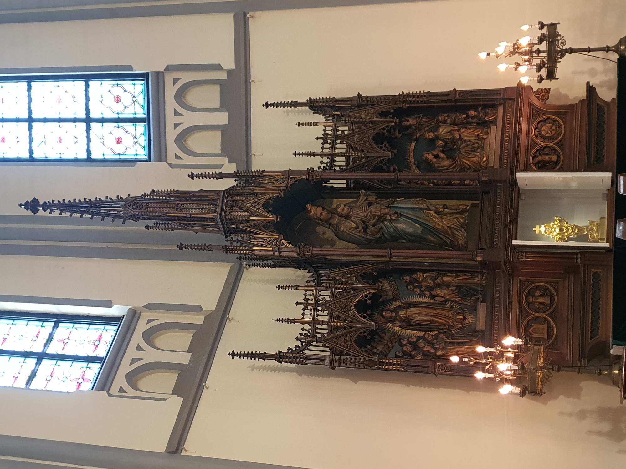 The image shows an ornate, dark wood altar with intricate carvings inside a church. The altar is dominated by a central sculpture of the Virgin Mary holding the infant Jesus, flanked by other religious figures. Above the altar are two stained-glass windows with floral patterns. To the left and right of the altar are candelabra with lit candles. The walls of the church are painted in light colors, and there are architectural details above the windows. In the lower center of the altar, a golden monstrance is visible within a recessed compartment. The scene appears to be from a solemn religious service. This scene is located in the Cathedral of Our Lady of Assumption, Jakarta, Indonesia.