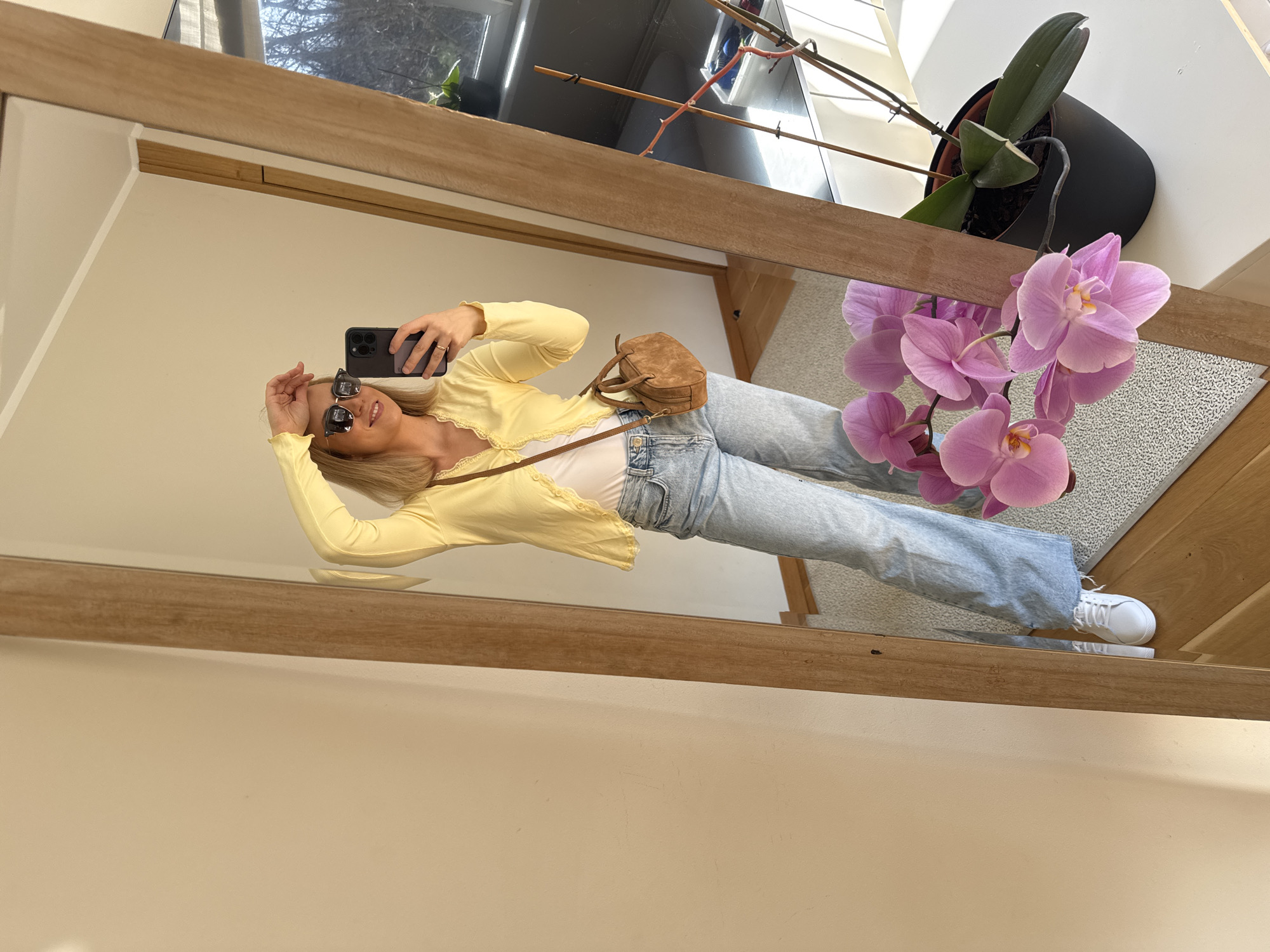 A woman is reflected in a mirror. She wears a yellow cardigan, light-wash jeans, and white sneakers. She is holding a smartphone to take a picture. There is a handbag on her shoulder. Above her reflection, a potted orchid with pink flowers is visible. Reflected behind her are glimpses of a window and another plant. The scene appears to be indoors. There is no way to determine from the image alone that the location is Riga, Latvia.