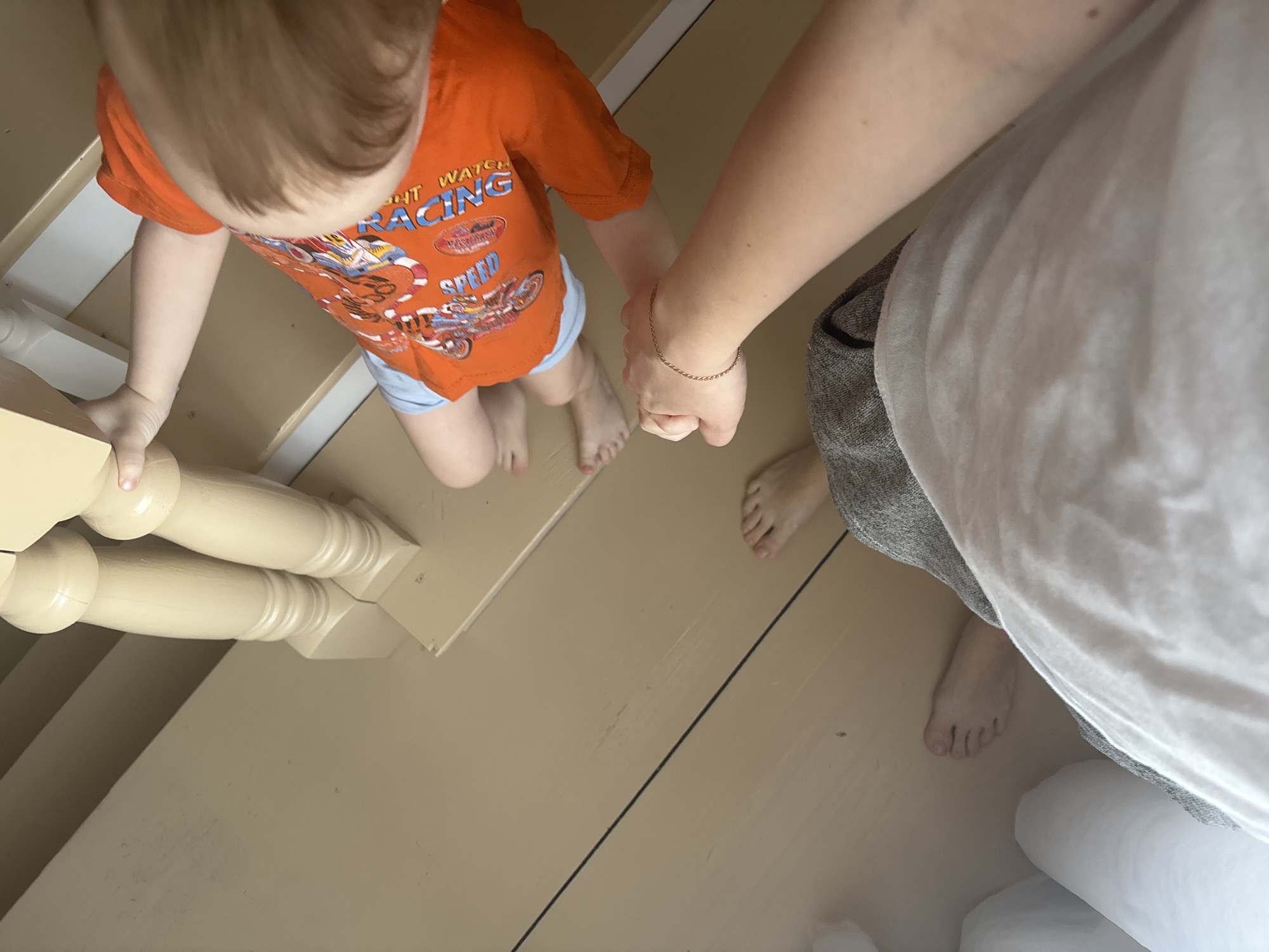 A top-down shot depicts a child wearing an orange "RACING" t-shirt and blue shorts descending wooden stairs, holding onto the railing and the hand of an adult. The adult, wearing grey shorts and a white shirt, is standing on the floor. The location is consistent with the type of domestic structure found in Ферма, Russia.