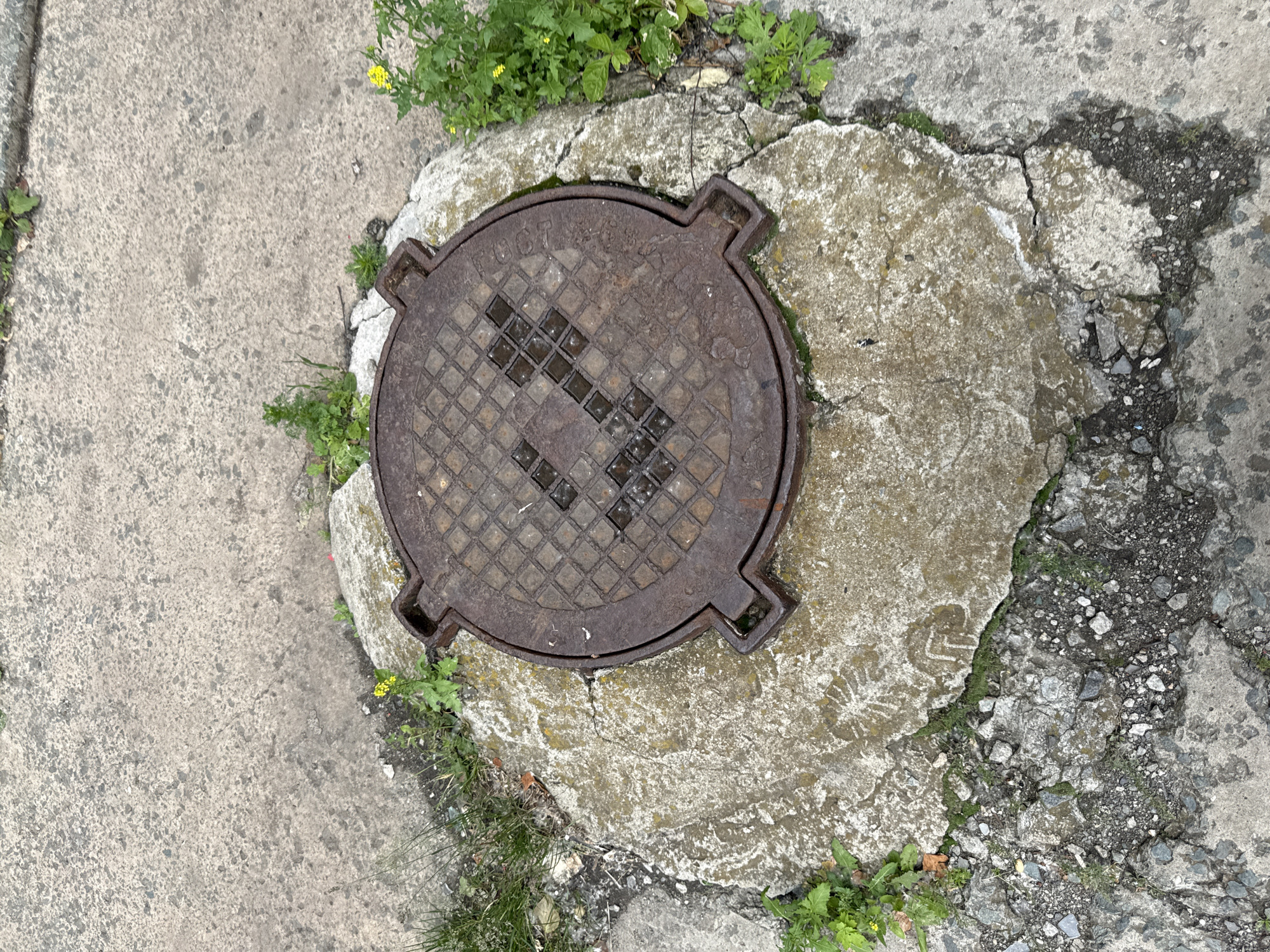 A rusty, circular metal manhole cover is centered on a cracked, irregular concrete slab that is surrounded by asphalt and overgrown weeds. The manhole cover has a textured, diamond-patterned surface with a stylized, dark-colored square or "check mark" symbol formed by four smaller squares within the larger pattern. The concrete slab is rough and weathered, with visible chips and cracks. Green weeds and some yellow wildflowers are growing around the edges of the concrete and in the cracks. The asphalt surface appears worn and textured, typical of a street or sidewalk. The scene suggests an outdoor urban environment. The image was captured at an oblique angle, looking down at the manhole cover. There are no people or animals visible in the image.