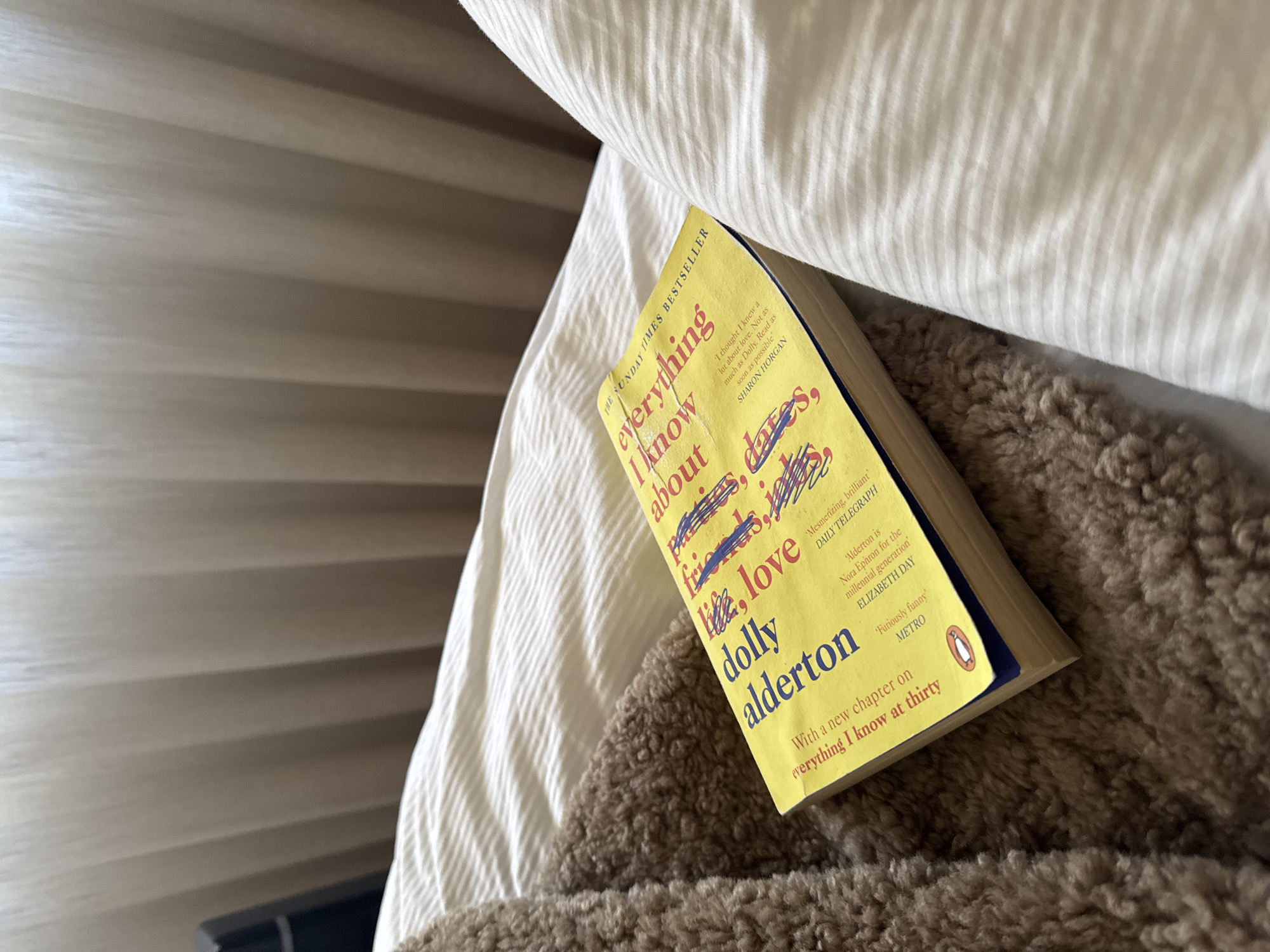 The image shows a paperback copy of Dolly Alderton's "Everything I Know About Love" resting on a bed. The book is yellow with blue and red scribbles on the cover. The bed has white striped sheets and a brown, furry throw. Part of a window blind is visible in the background.