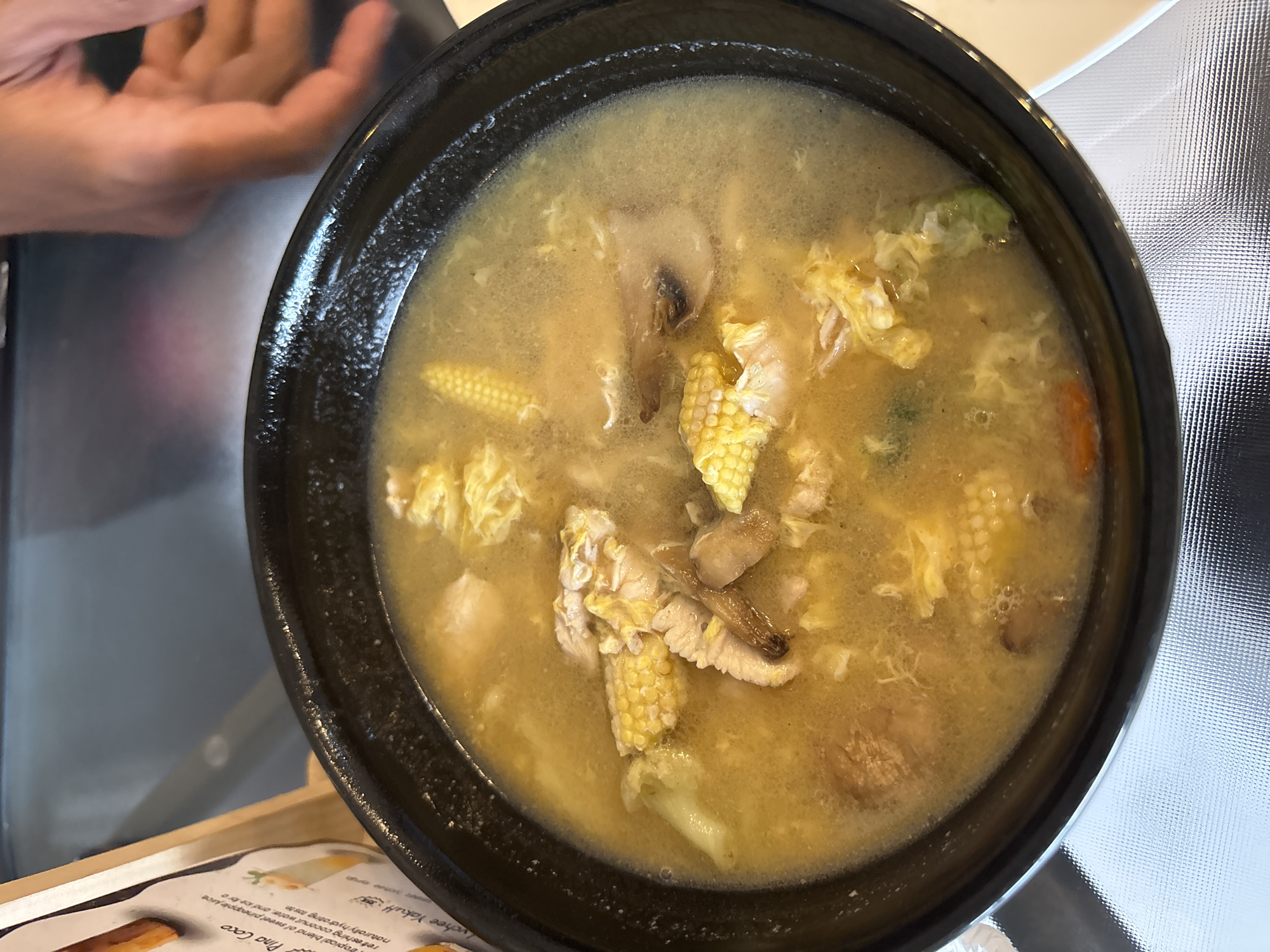 A close-up, overhead shot shows a bowl of soup with corn, mushrooms, and shredded egg. A hand is visible in the upper right corner, reaching towards the bowl. The soup is served in a black bowl, placed on a reflective surface. In the background, a menu board with text in English and an image of a drink is propped up. The overall scene appears to be within a restaurant or food establishment, likely in Kuta Selatan, Indonesia, given the context.